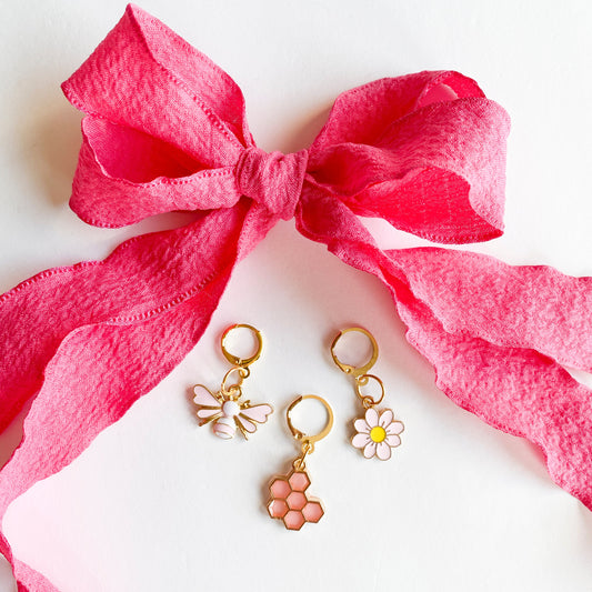 Gold shoe charms with pink bee, honeycomb, and white daisy designs, displayed with bright pink ribbon shoelaces tied in a bow on white background.