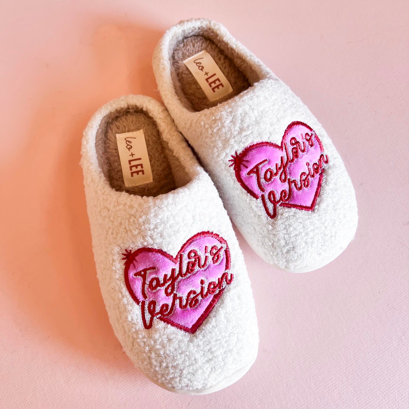 Pair of cozy slippers from Taylor's Version collection, featuring a soft, plush exterior in a light beige color with embroidered detailing and a fluffy interior lining for maximum comfort