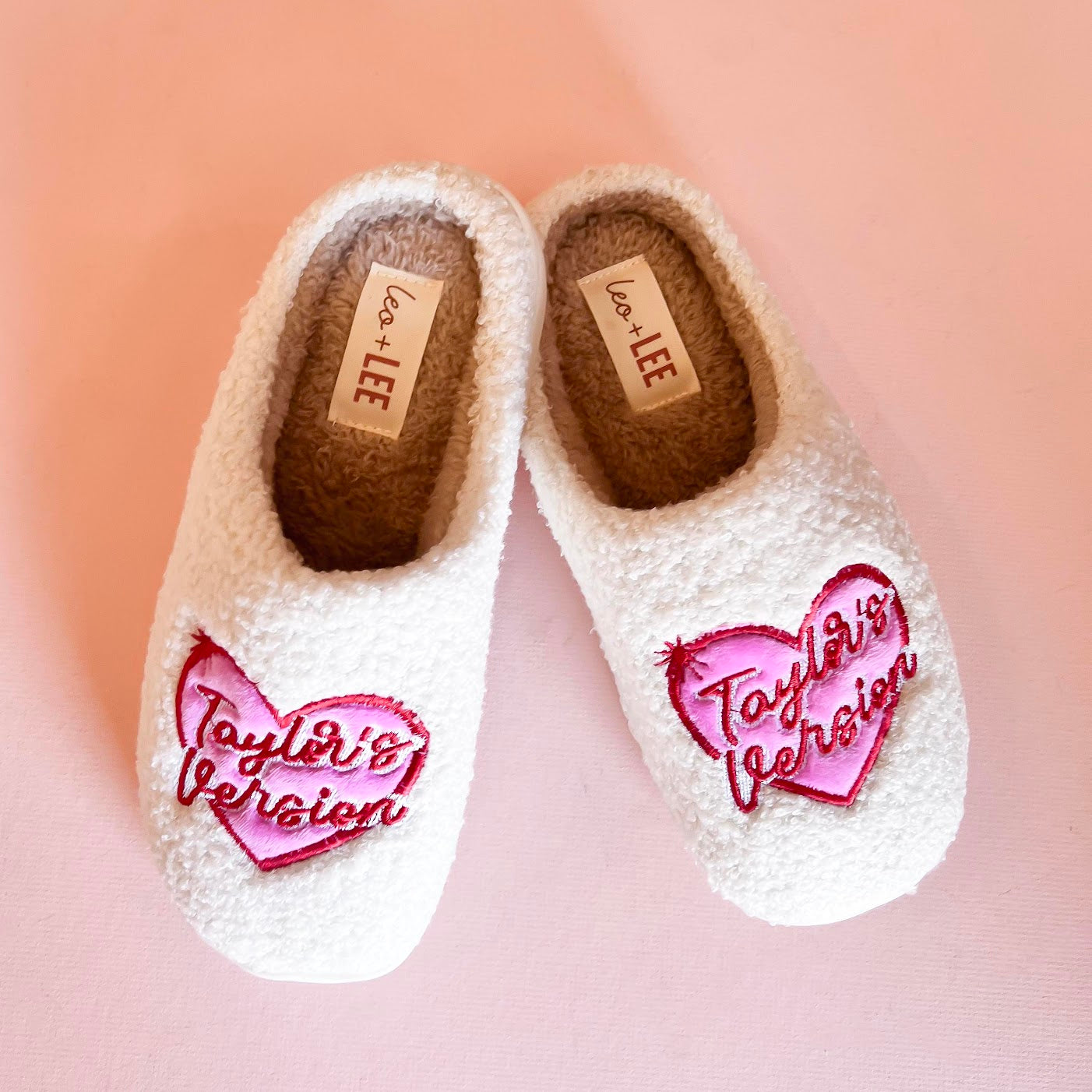Pair of cozy slippers from Taylor's Version collection, featuring a soft, plush exterior in a light beige color with embroidered detailing and a fluffy interior lining for maximum comfort