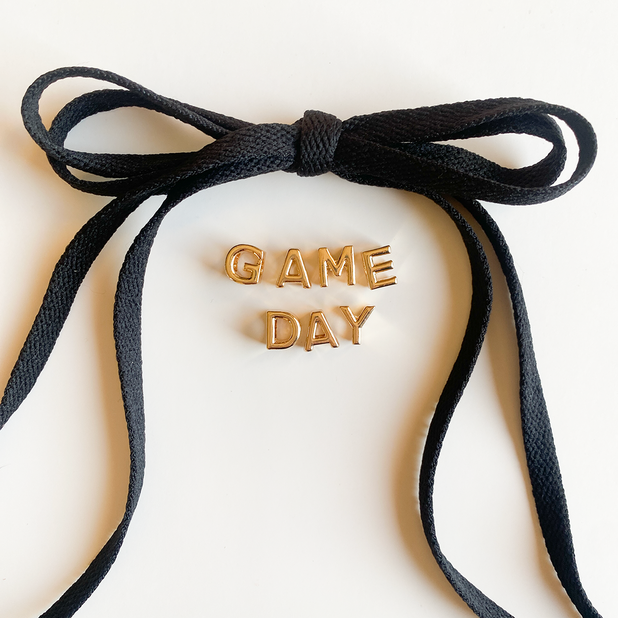 A close-up image of rich black shoelaces tied in a bow, accompanied by gold letter charms spelling "GAME DAY" displayed below.