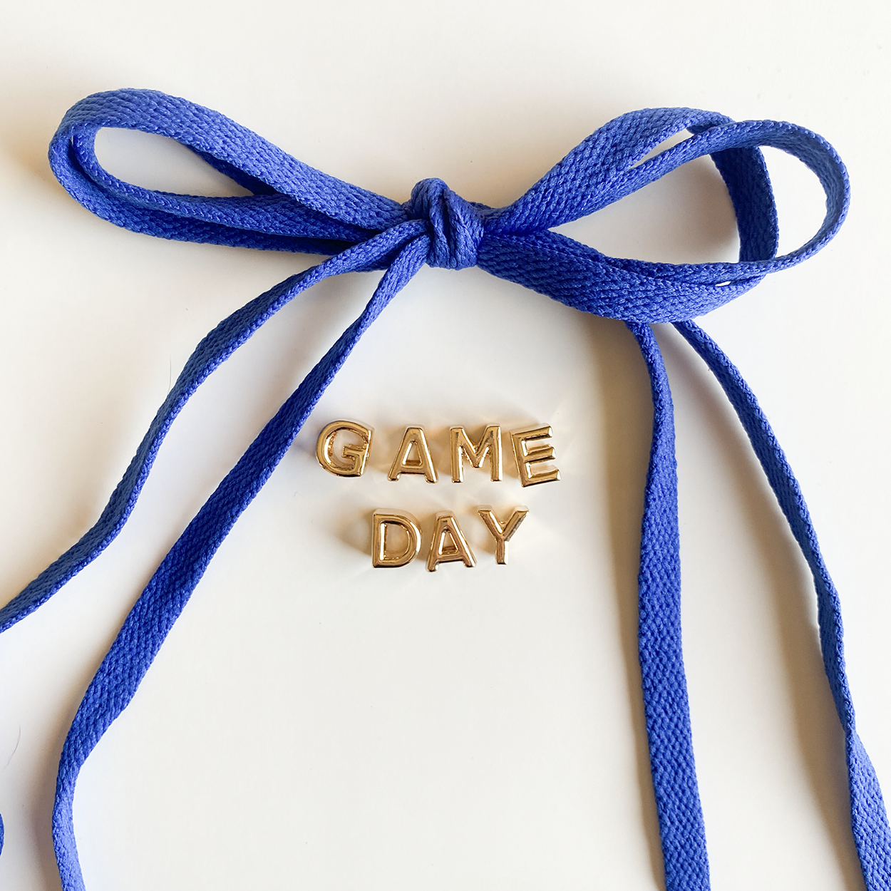A close-up image of rich blue shoelaces tied in a bow, accompanied by gold letter charms spelling "GAME DAY" displayed below.