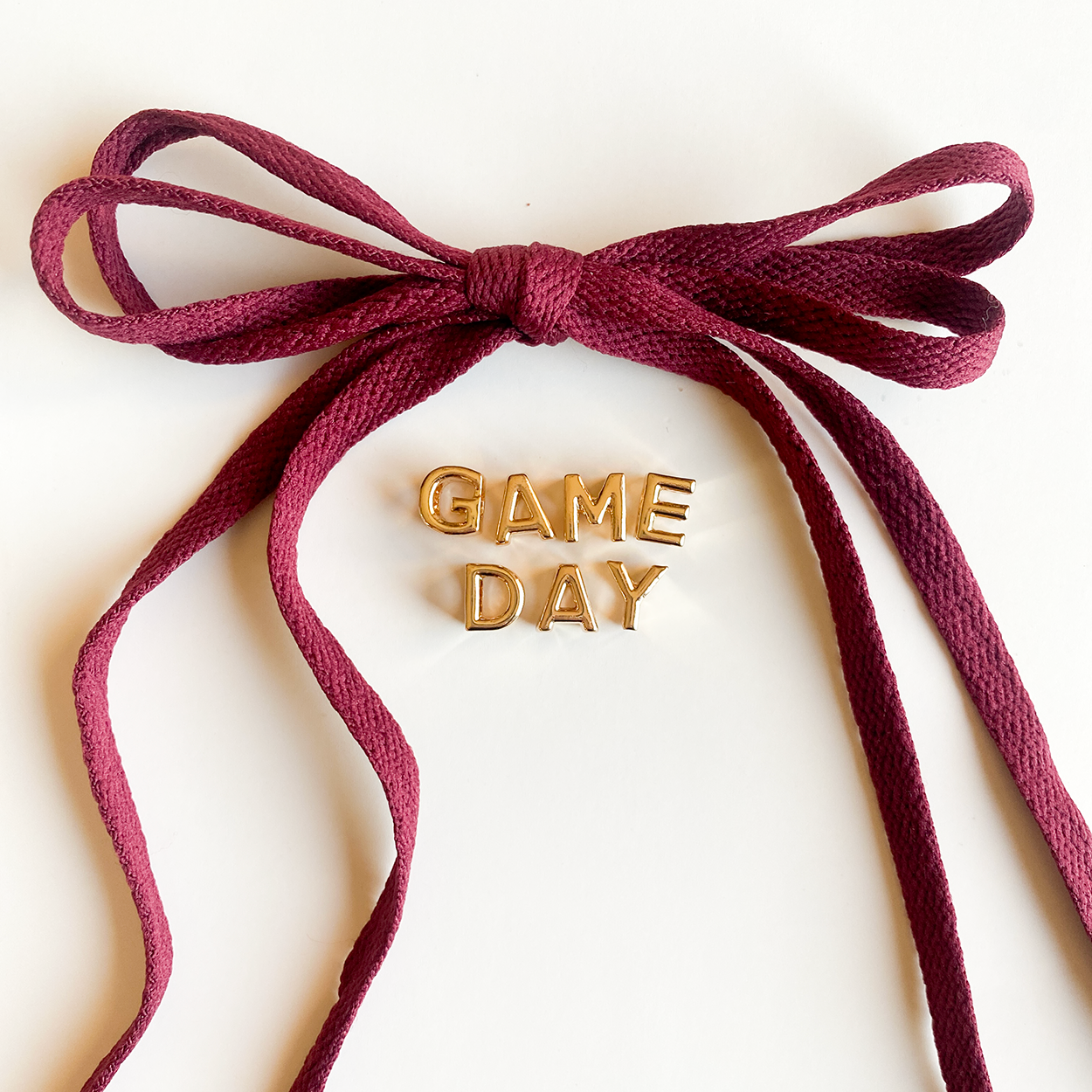 A close-up image of rich crimson shoelaces tied in a bow, accompanied by gold letter charms spelling "GAME DAY" displayed below.