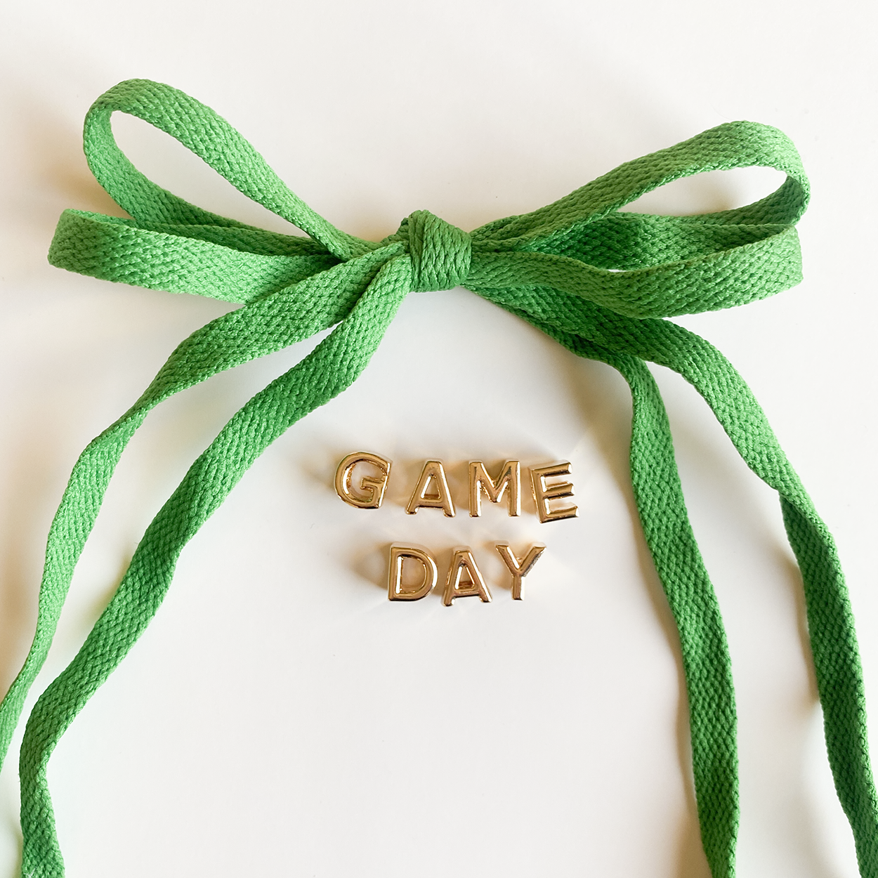 A close-up image of rich kelly green shoelaces tied in a bow, accompanied by gold letter charms spelling "GAME DAY" displayed below.