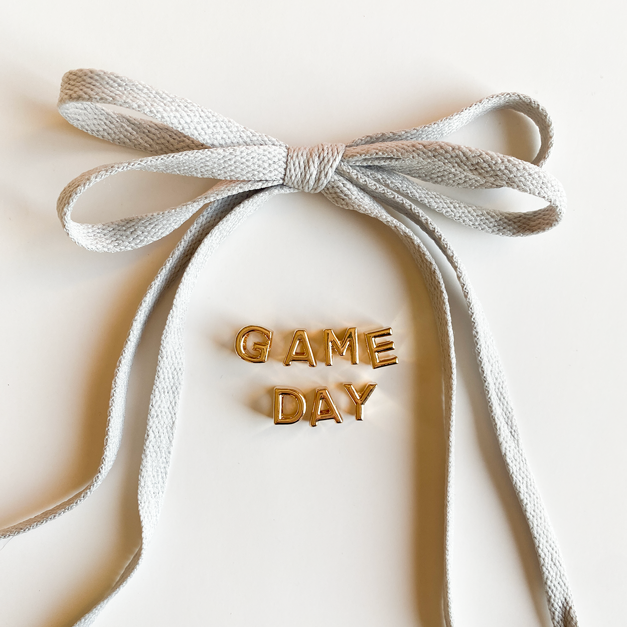 A close-up image of rich grey shoelaces tied in a bow, accompanied by gold letter charms spelling "GAME DAY" displayed below.