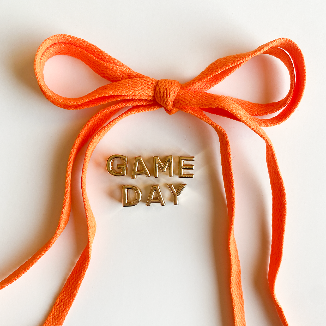 A close-up image of rich orange shoelaces tied in a bow, accompanied by gold letter charms spelling "GAME DAY" displayed below.