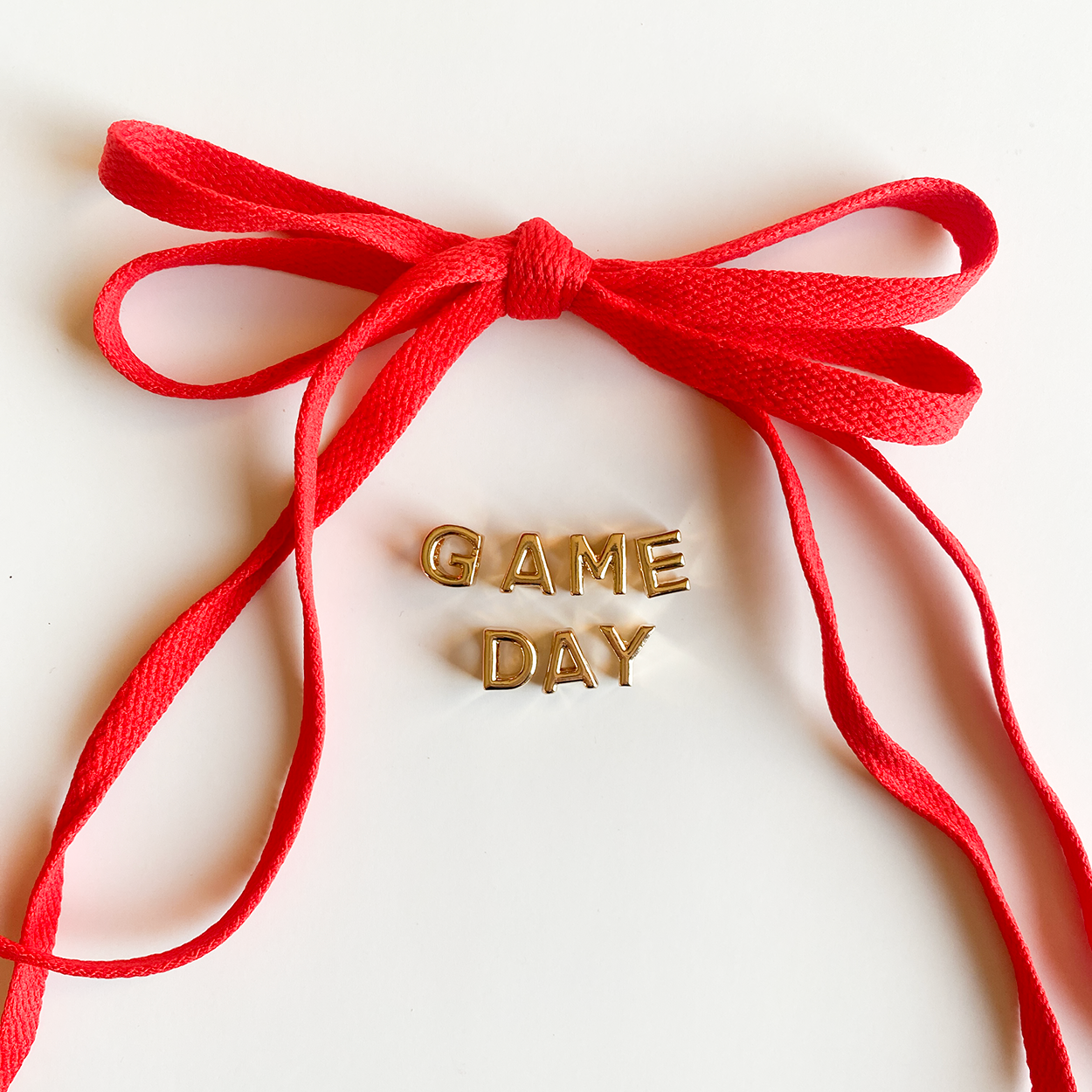 A close-up image of rich red shoelaces tied in a bow, accompanied by gold letter charms spelling "GAME DAY" displayed below.