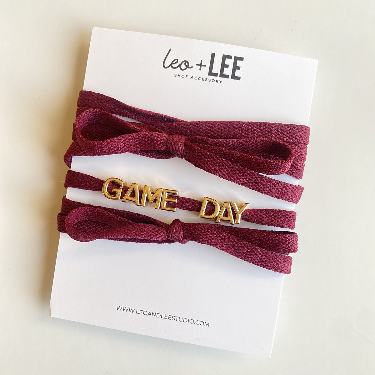 A close-up image of rich crimson shoelaces tied in a bow, accompanied by gold letter charms spelling "GAME DAY" displayed below.