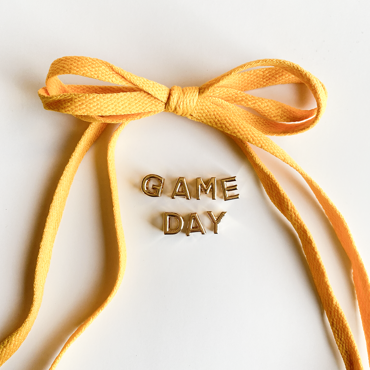 A close-up image of rich golden yellow shoelaces tied in a bow, accompanied by gold letter charms spelling "GAME DAY" displayed below.