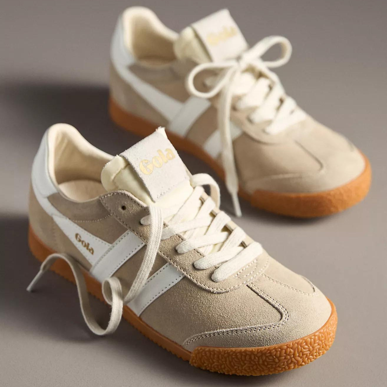 A pair of Gola Elan sneakers in a bone and white colorway, featuring a suede upper with white leather stripes, cream laces, and a gum rubber sole. The "Gola" logo is embossed in gold on the tongue and side.