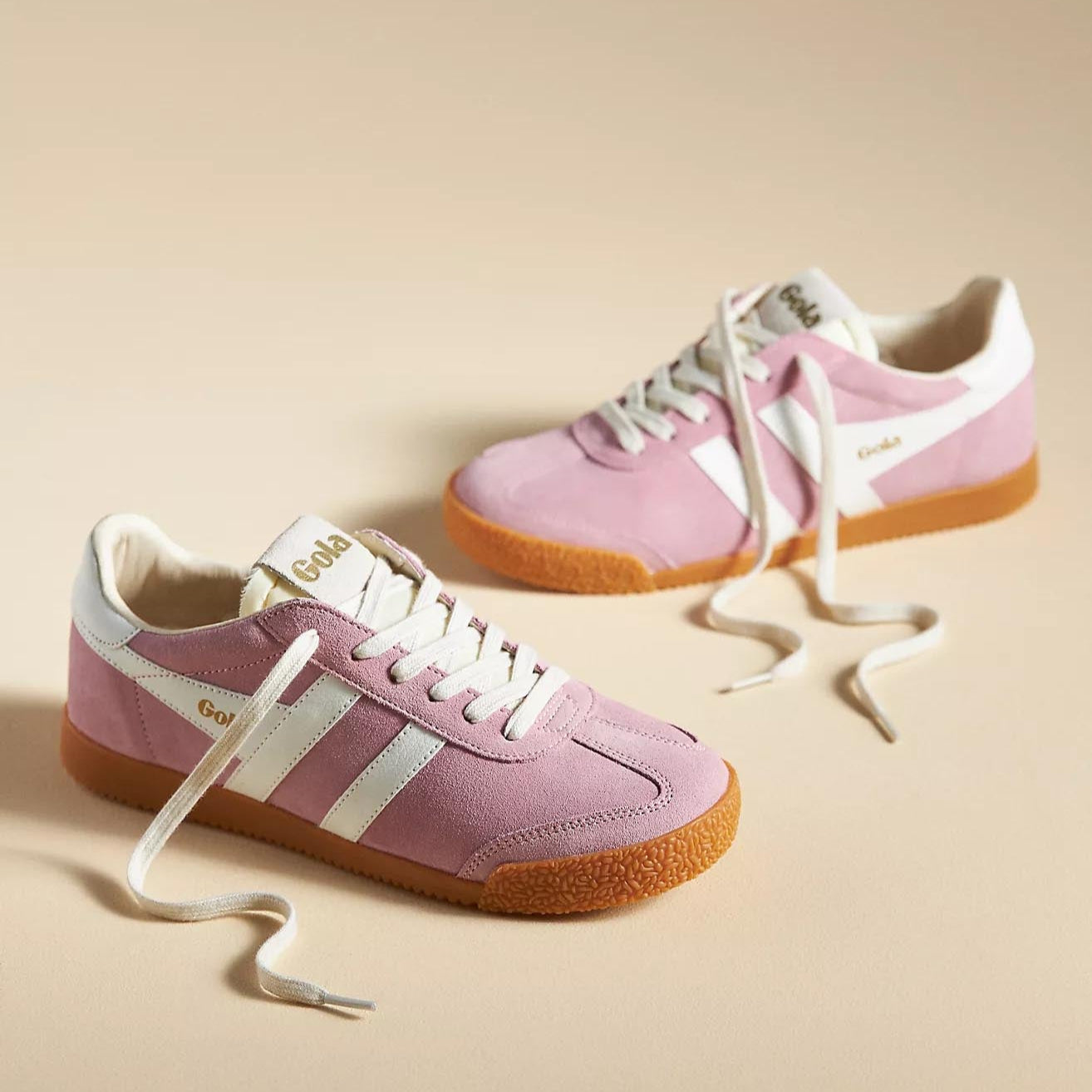 A close-up of the Gola Elan sneakers in Pink Candy & White, featuring a soft pink suede upper, white leather stripes, and a gum rubber sole. The "Gola" logo is embossed in gold on the side, with cream laces adding to the retro aesthetic.