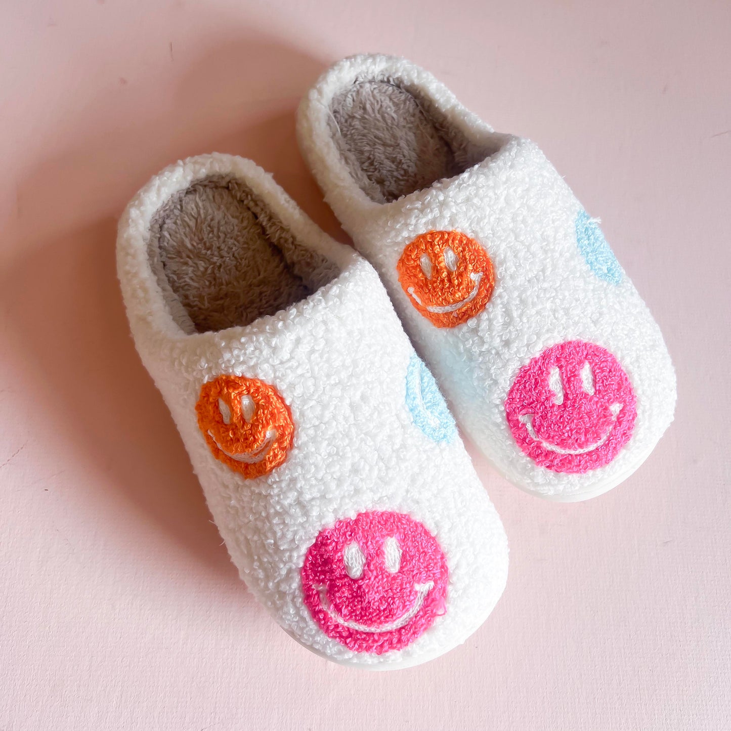 Fluffy white slippers adorned with colorful embroidered smiley faces in pink, orange, and blue. These cozy and fun slippers are perfect for adding a touch of happiness and comfort to your indoor footwear collection.