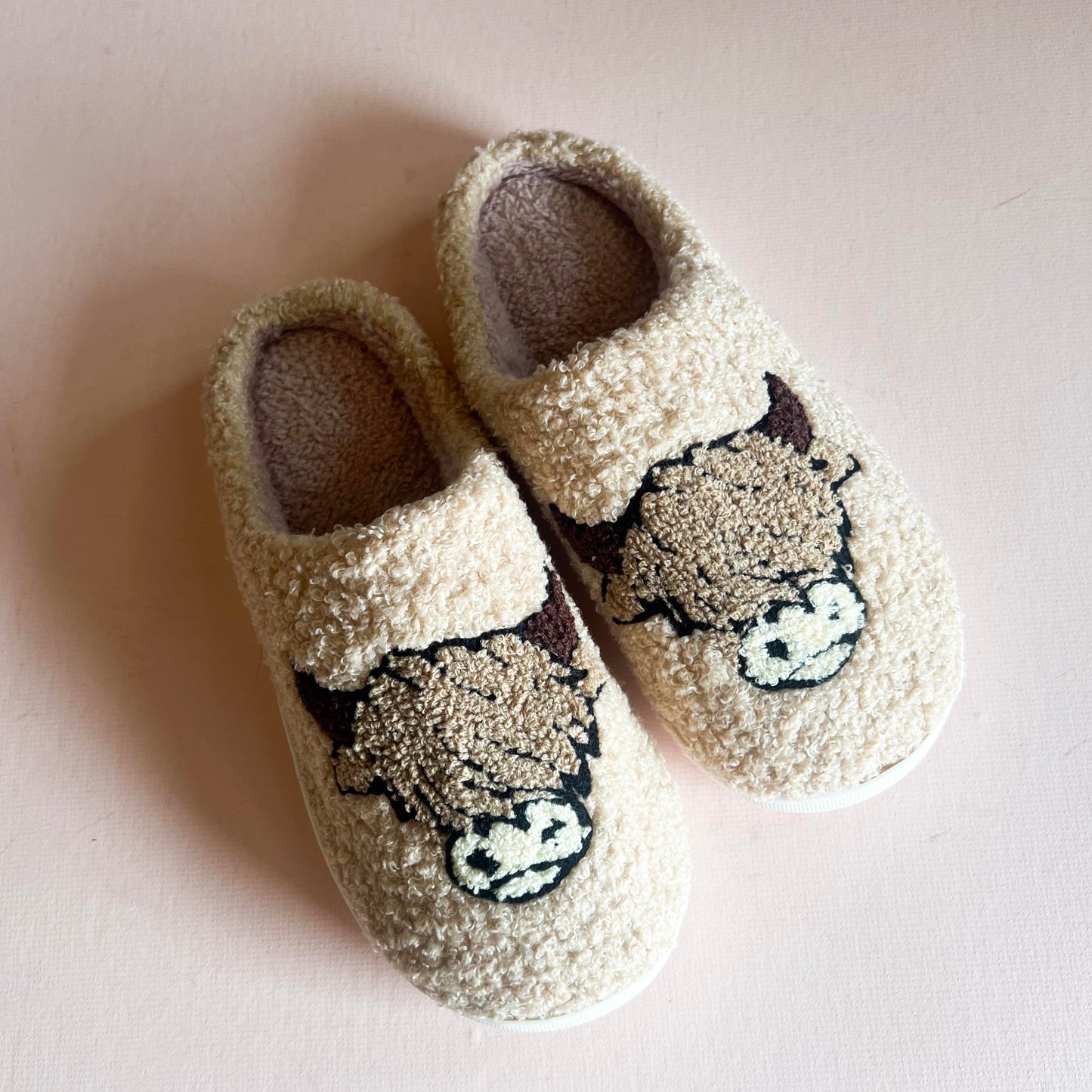 Cozy and fluffy beige slippers featuring an adorable embroidered highland cow design on the top. Perfect for keeping your feet warm and comfortable at home.