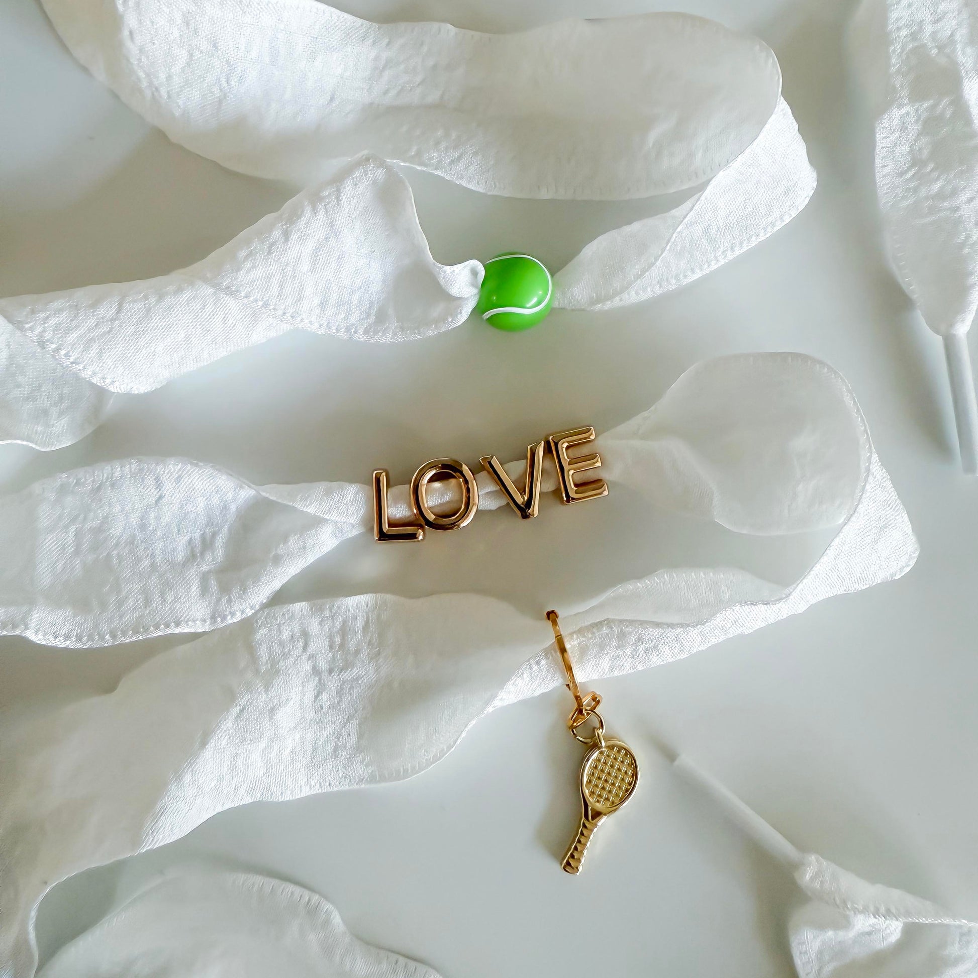 Flat lay of Leo & Lee tennis sneaker charm set with a green tennis ball charm, gold LOVE charm, and gold tennis racquet charm on white ribbon shoelaces.