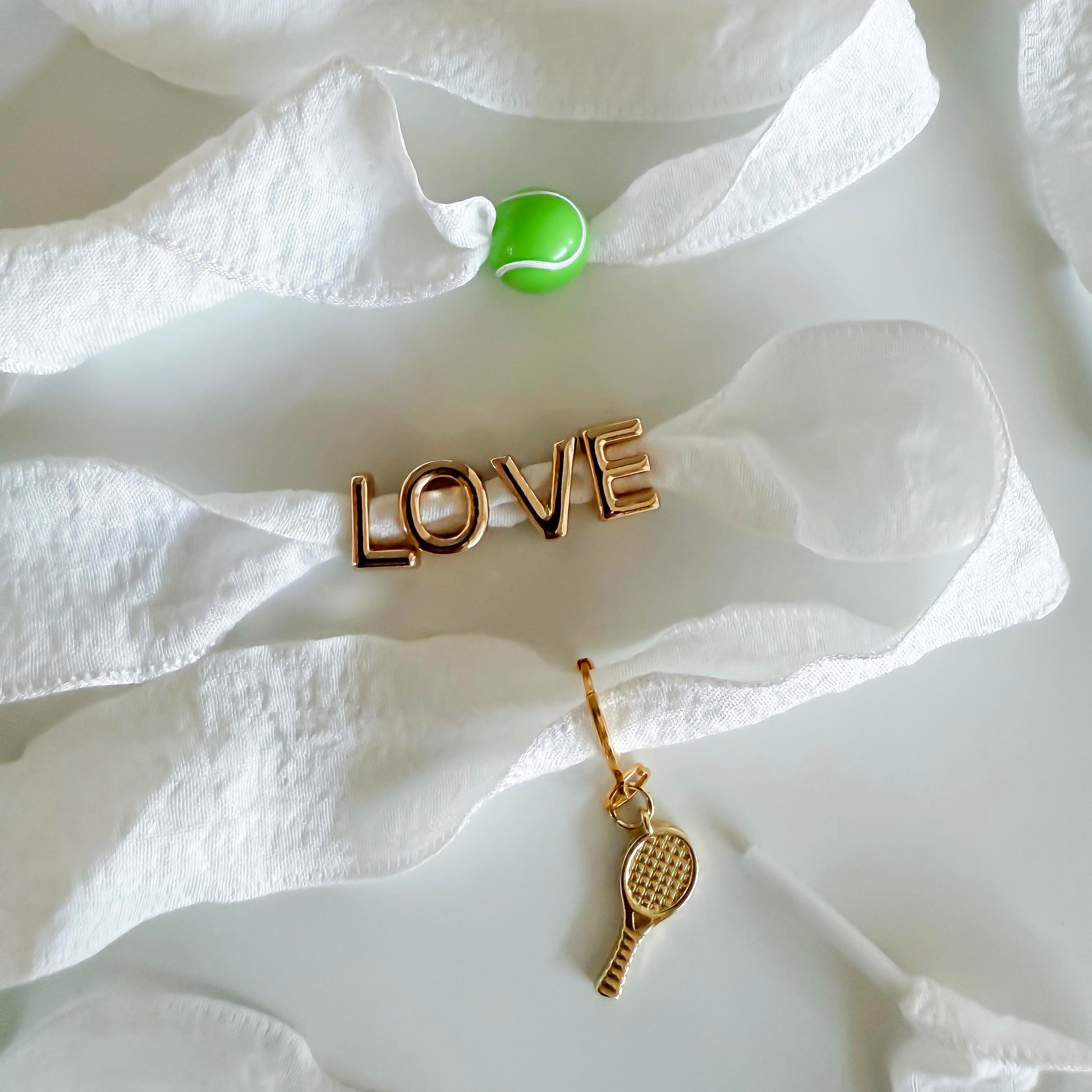 White sneaker styled with Leo & Lee tennis-themed sneaker charms: green tennis ball, gold LOVE charm, and dangling gold racquet on wide ribbon laces.