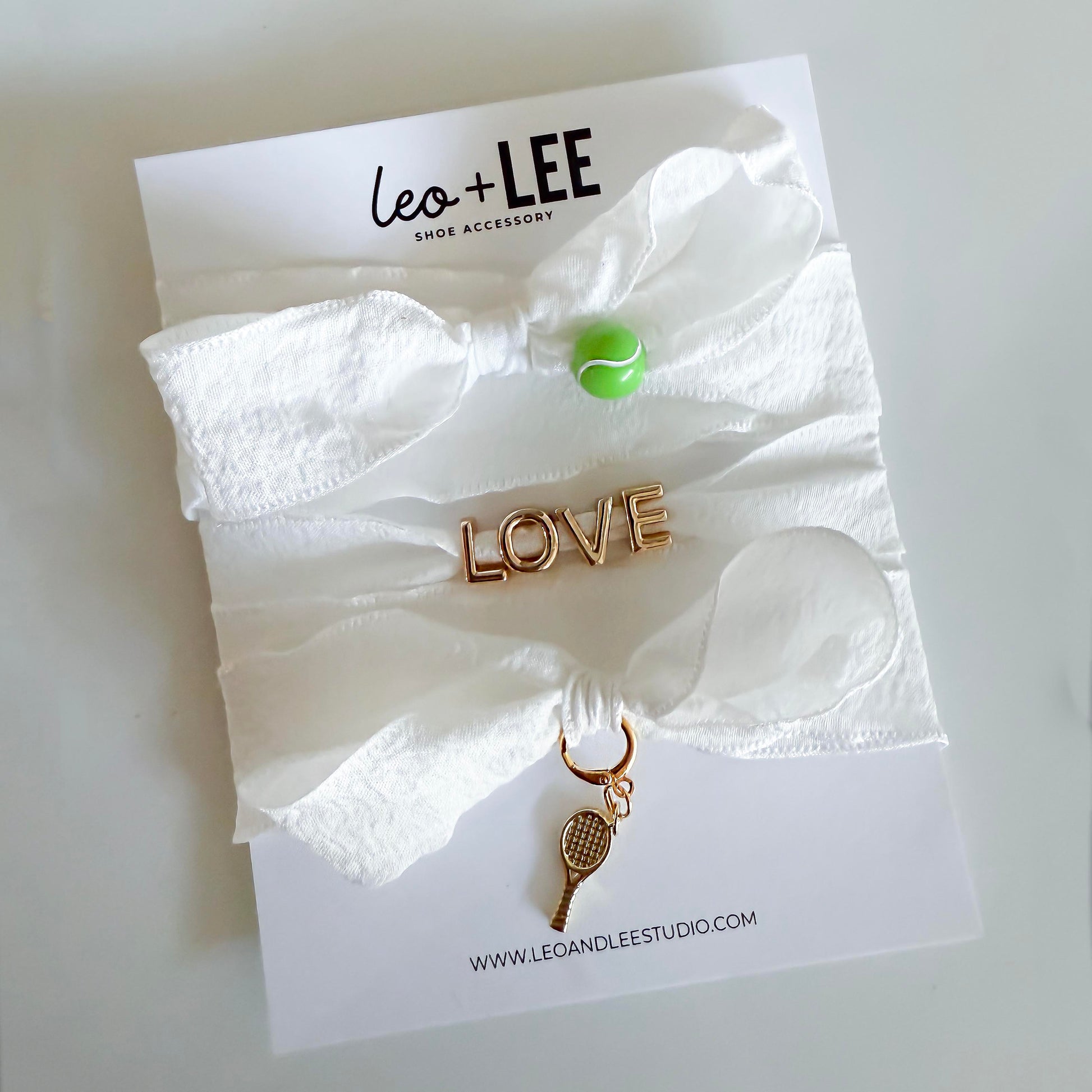 Flat lay of Leo & Lee tennis sneaker charm set with a green tennis ball charm, gold LOVE charm, and gold tennis racquet charm on white ribbon shoelaces.