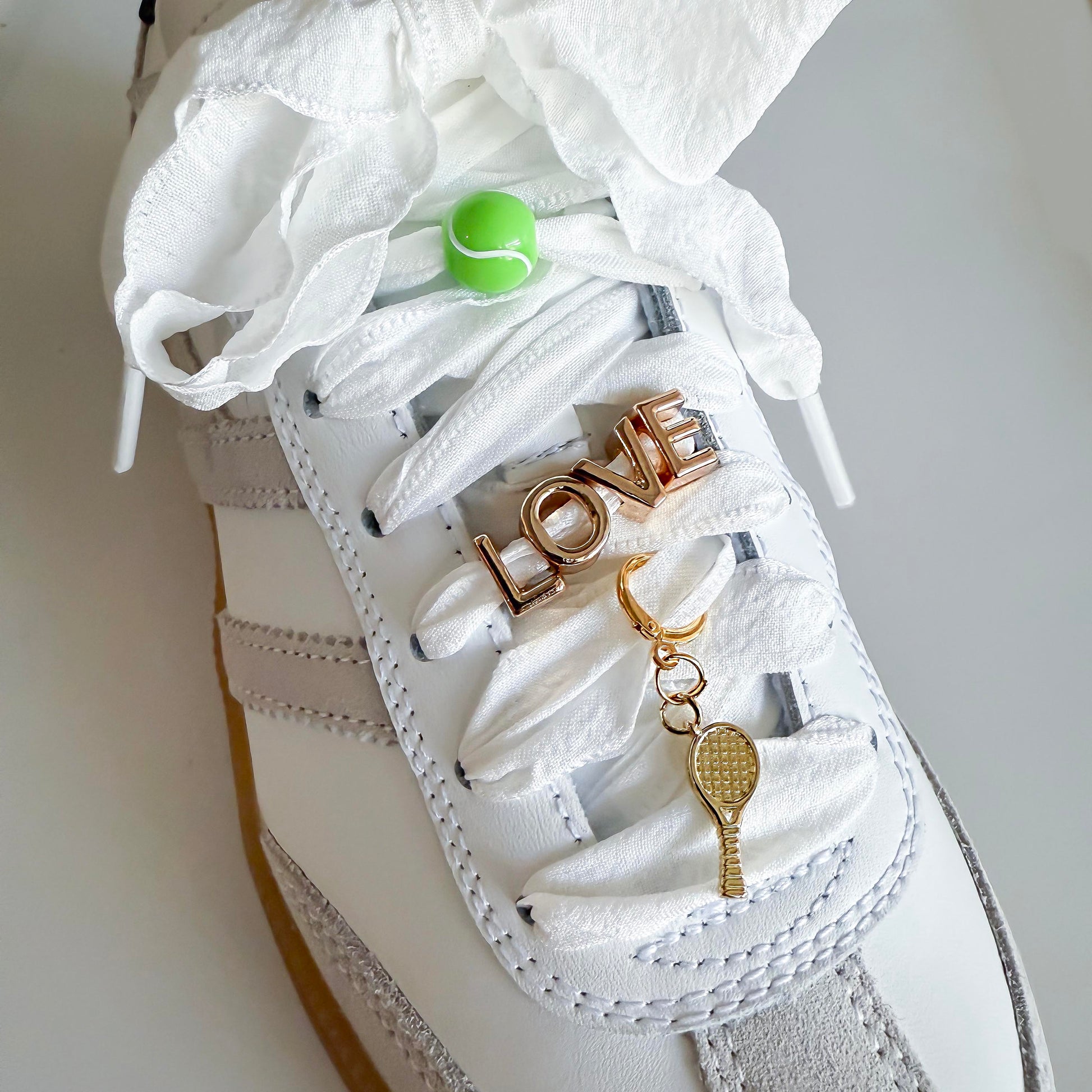 White sneaker styled with Leo & Lee tennis-themed sneaker charms: green tennis ball, gold LOVE charm, and dangling gold racquet on wide ribbon laces.