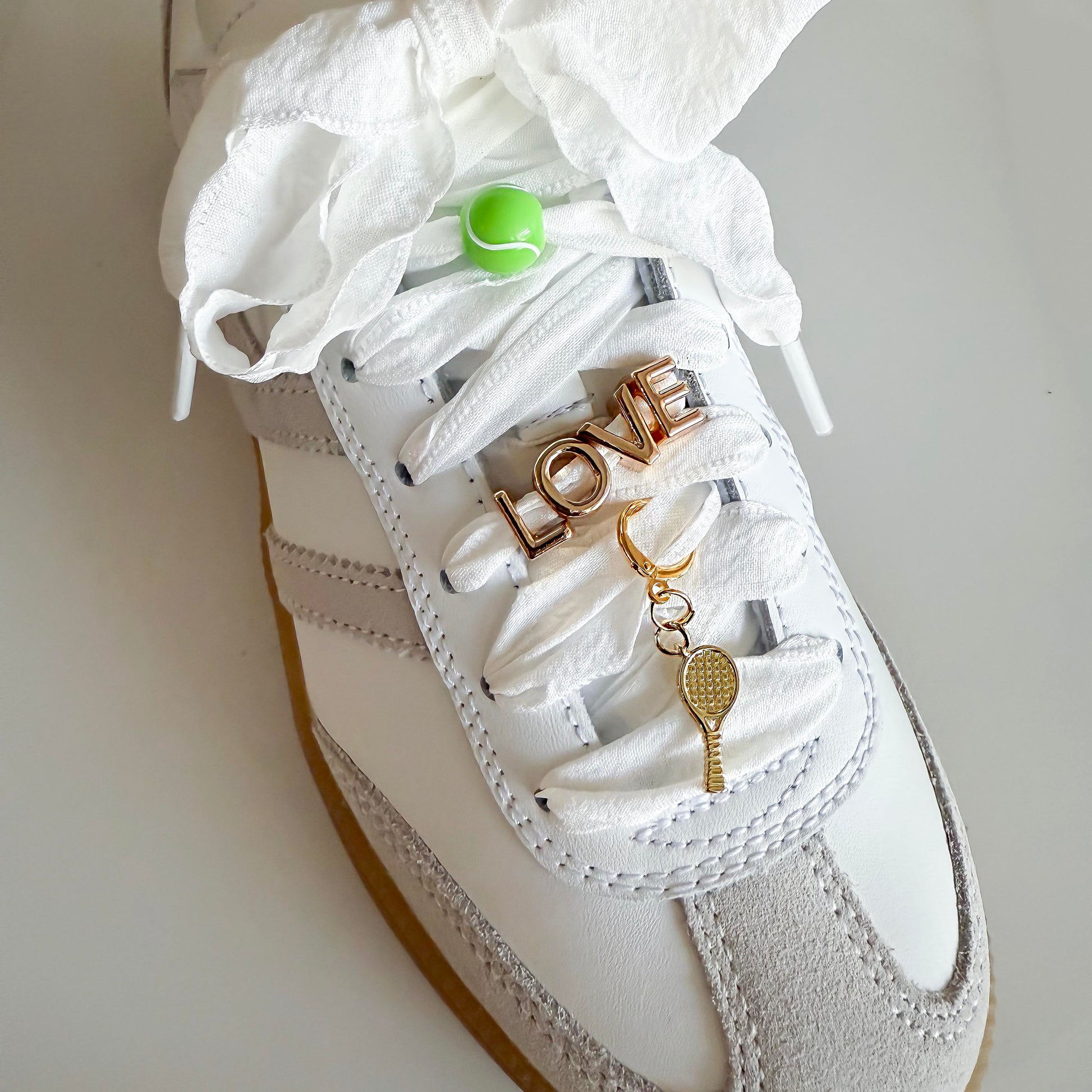 White sneaker styled with Leo & Lee tennis-themed sneaker charms: green tennis ball, gold LOVE charm, and dangling gold racquet on wide ribbon laces.