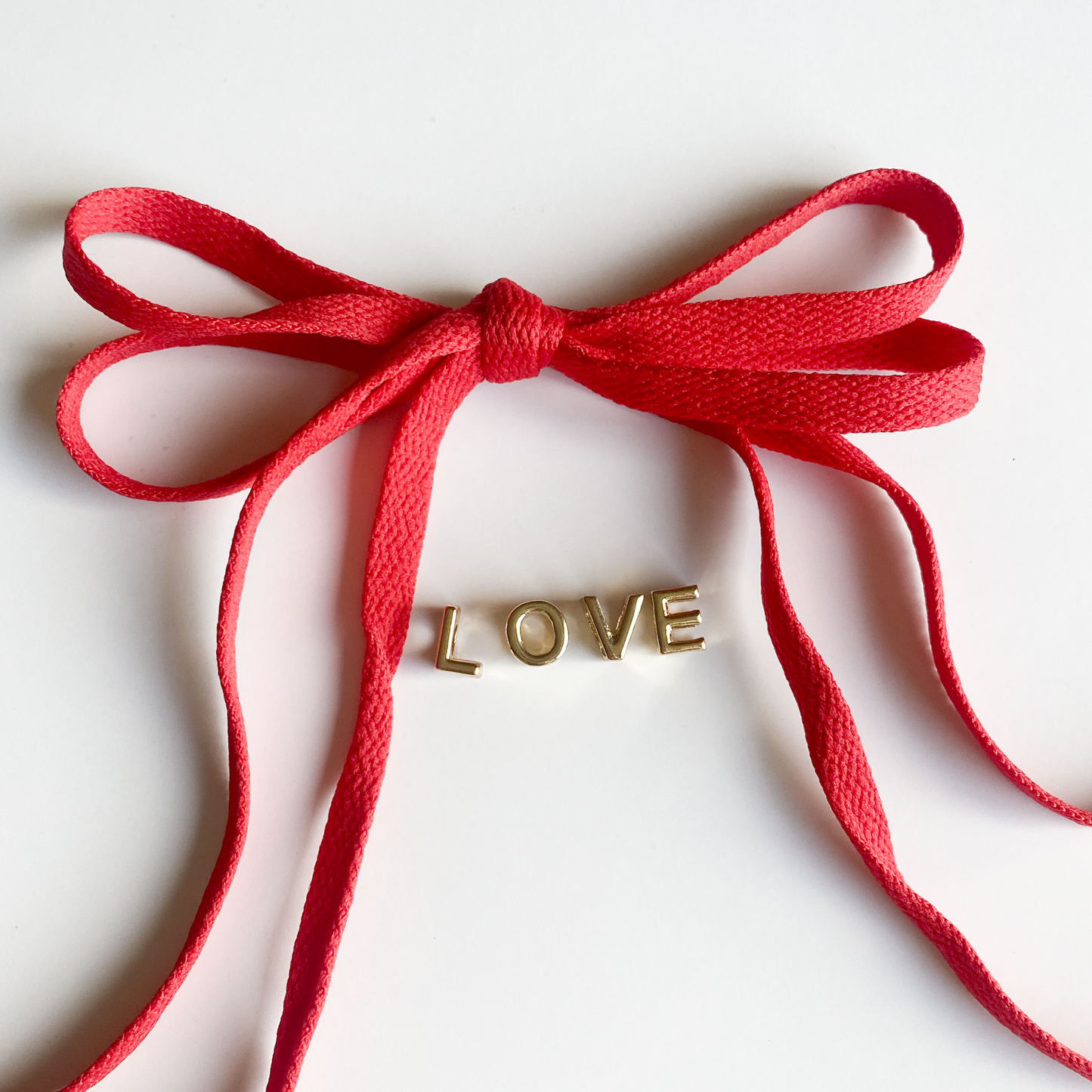 Red and white bow shoelaces with gold 'LOVE' charms from the Leo + Lee Sneaker Charm Set, displayed in packaging and styled individually.