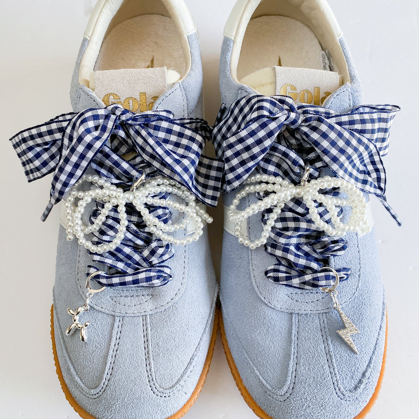 A set of navy gingham ribbon shoe laces and pearl strand laces displayed on a Leo + Lee card, featuring two silver charms — a balloon dog and a rhinestone lightning bolt — arranged neatly on a white background.