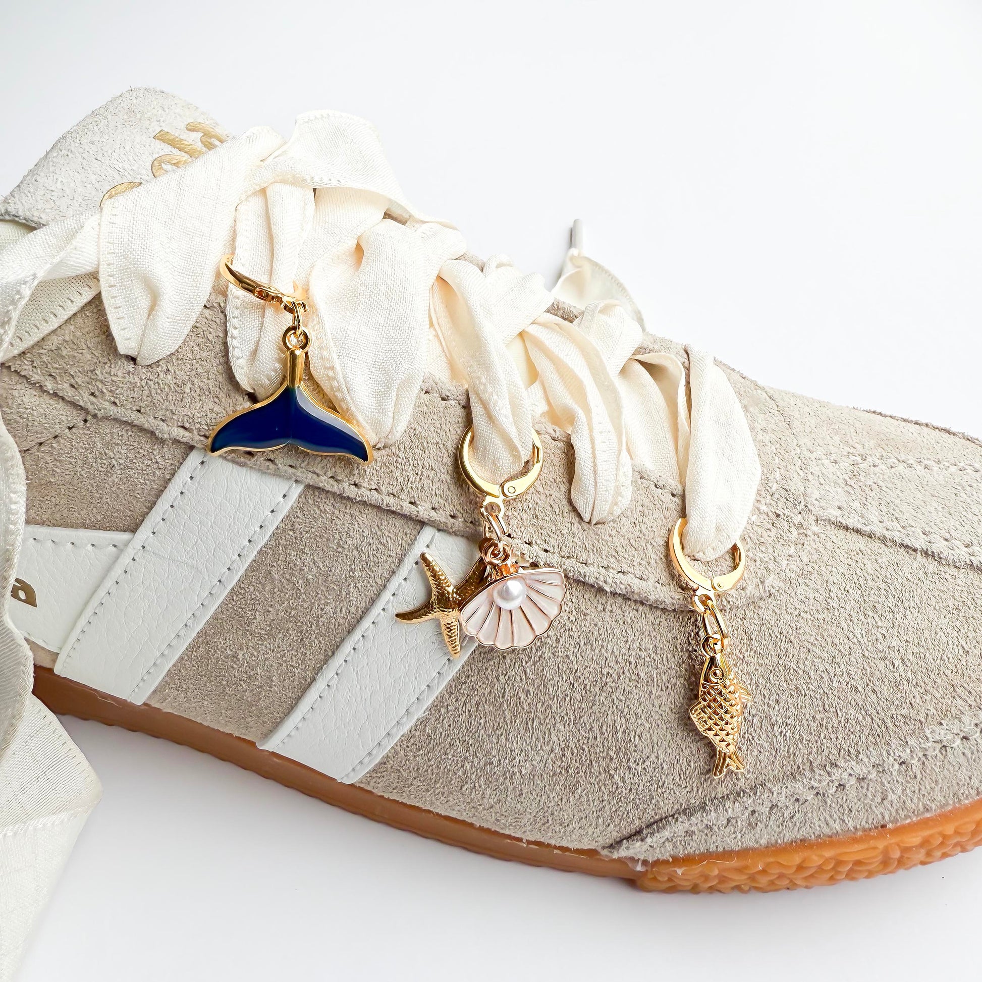Beige sneaker with white satin laces featuring gold ocean-themed shoelace charms including a whale tail, starfish, seashell, and conch shell, styled flat on a white background.