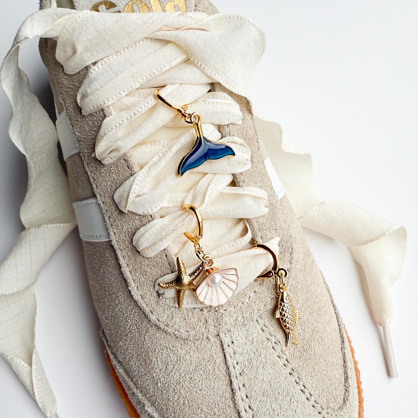Beige sneaker with white satin laces featuring gold ocean-themed shoelace charms including a whale tail, starfish, seashell, and conch shell, styled flat on a white background.