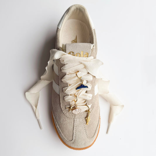 Beige sneaker with white satin laces featuring gold ocean-themed shoelace charms including a whale tail, starfish, seashell, and conch shell, styled flat on a white background.