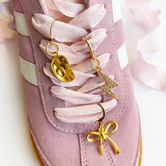 Light pink ribbon bow displayed with three gold sneaker charms — cowboy hat, lightning bolt with rhinestones, and bow — on a white background.