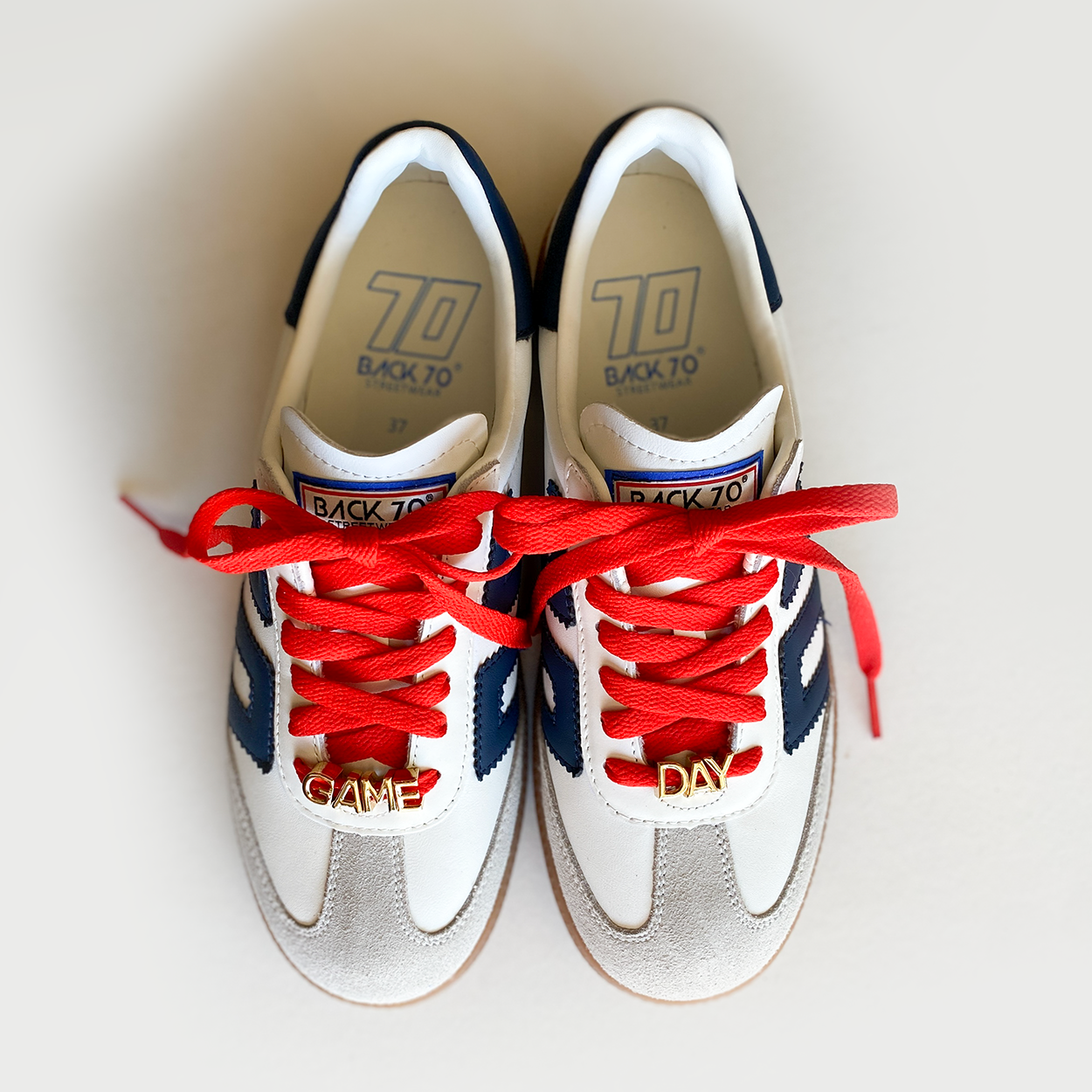 A close-up image of red shoelaces tied in a bow on a Back 70 sneaker accompanied by gold letter charms spelling "GAME DAY" displayed below.