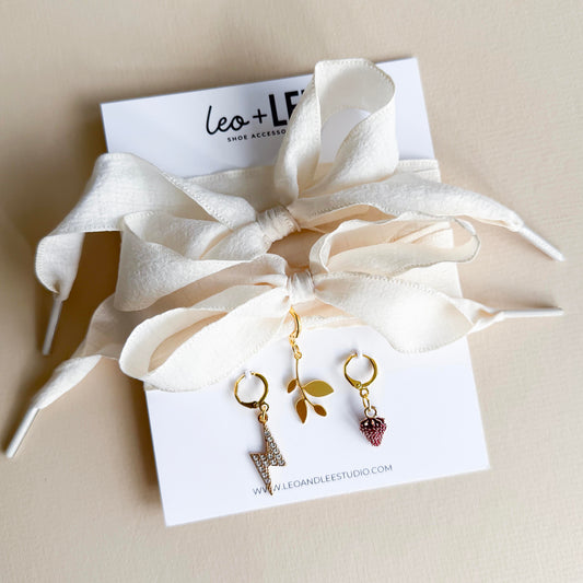 White ribbon shoelaces with gold sneaker charms – lightning bolt, leaf, and strawberry – displayed on product card by Leo + Lee Studio