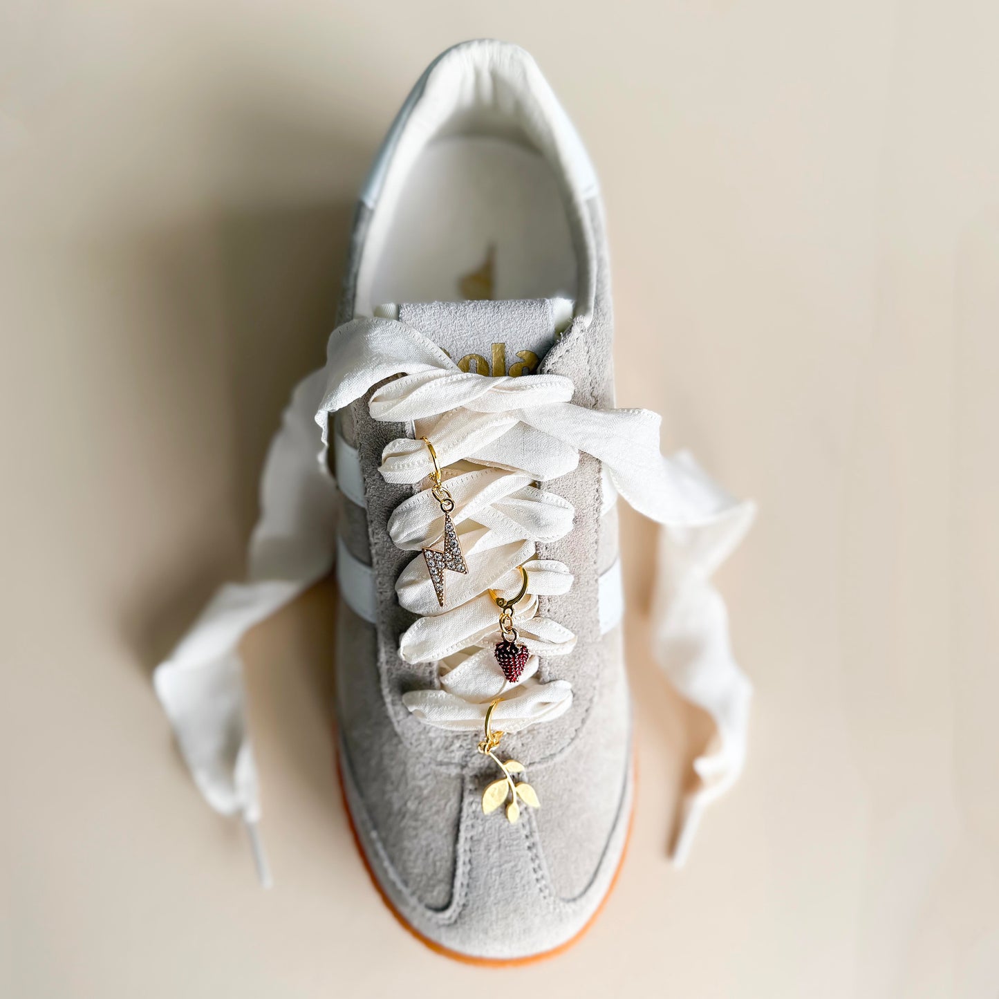 White ribbon shoelaces with gold sneaker charms – lightning bolt, leaf, and strawberry – displayed on product card by Leo + Lee Studio