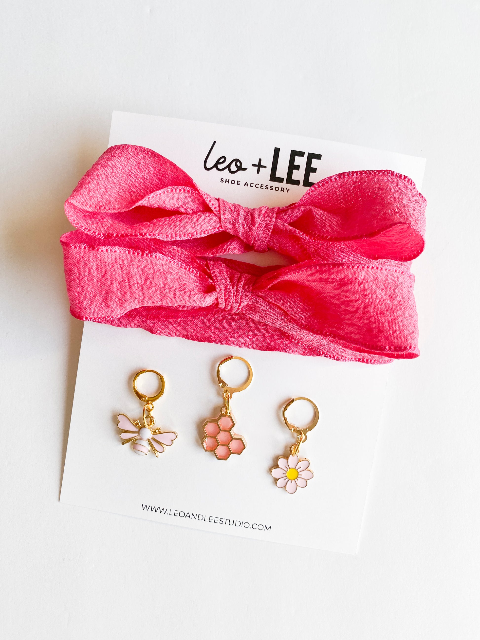 Gold shoe charms with pink bee, honeycomb, and white daisy designs, displayed with bright pink ribbon shoelaces tied in a bow on white background.