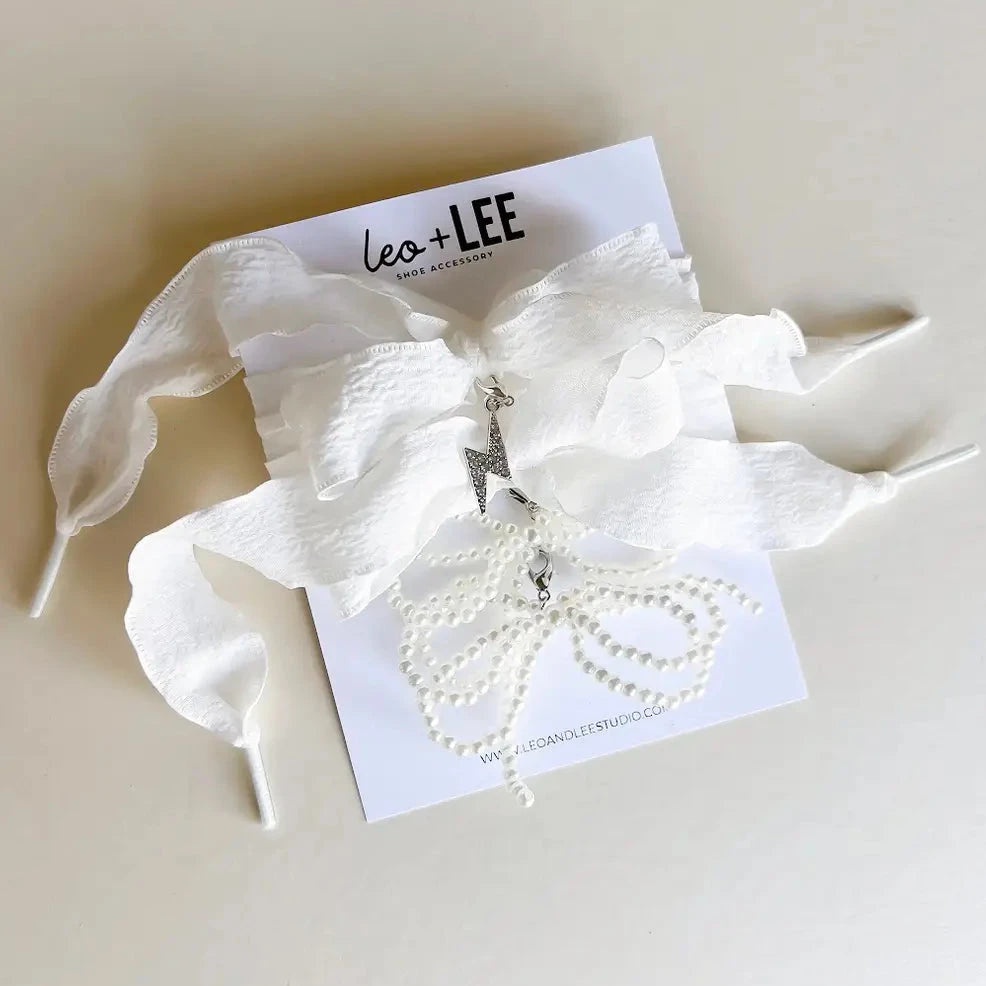 White shoe lace accessory with a lightning bolt charm and pearl bow charm on a branded card, set against a light background.