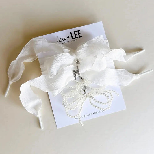 White shoe lace accessory with a lightning bolt charm and pearl bow charm on a branded card, set against a light background.