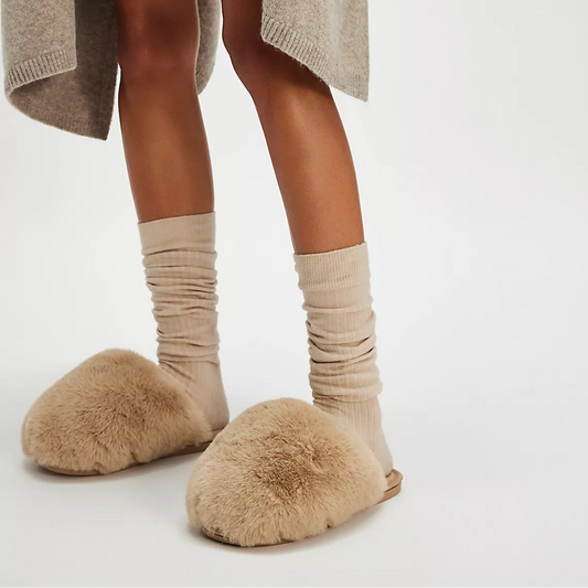 Person wearing a long beige cardigan, black dress with floral embroidery, beige socks, and fluffy slippers on a white background.