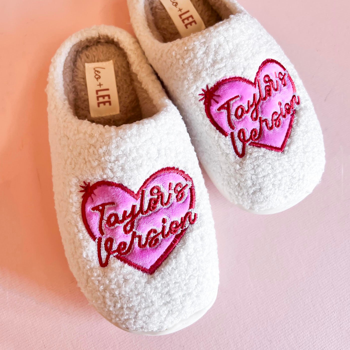 Pair of cozy slippers from Taylor's Version collection, featuring a soft, plush exterior in a light beige color with embroidered detailing and a fluffy interior lining for maximum comfort