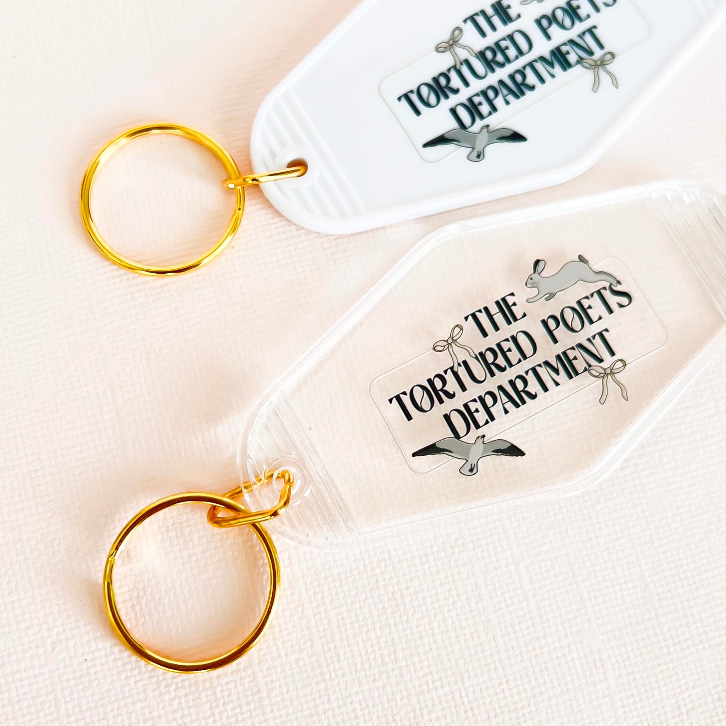 "Two vintage-inspired keychains labeled 'The Tortured Poets Department.' One is white with a golden keyring, and the other is clear with a golden keyring. Both feature a unique, playful design perfect for literary enthusiasts."