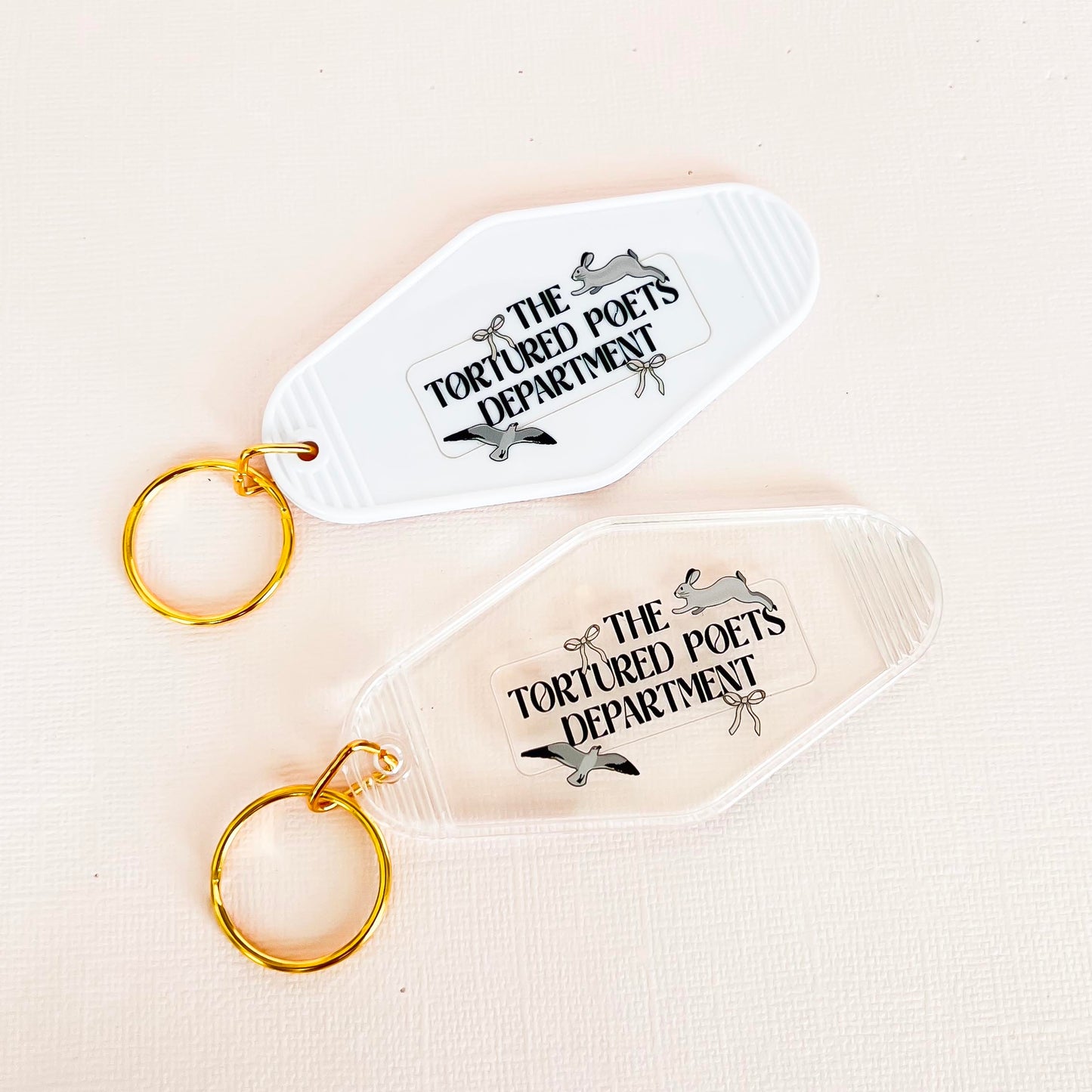 "Two vintage-inspired keychains labeled 'The Tortured Poets Department.' One is white with a golden keyring, and the other is clear with a golden keyring. Both feature a unique, playful design perfect for literary enthusiasts."