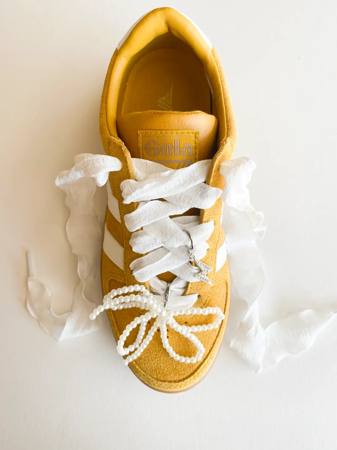 White shoe lace accessory with a lightning bolt charm and pearl bow charm on a branded card, set against a light background.