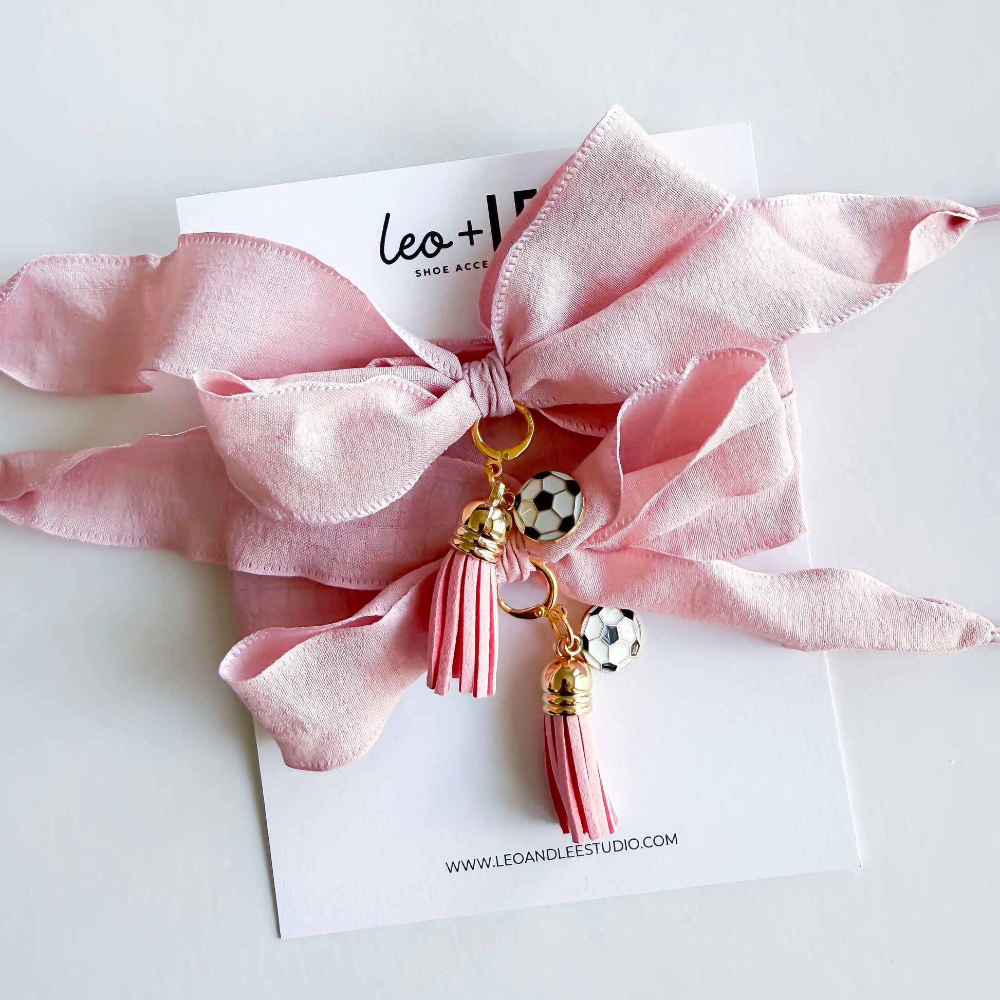 Pink silky bow laces with tassels and soccer charms on a white card with brand name.
