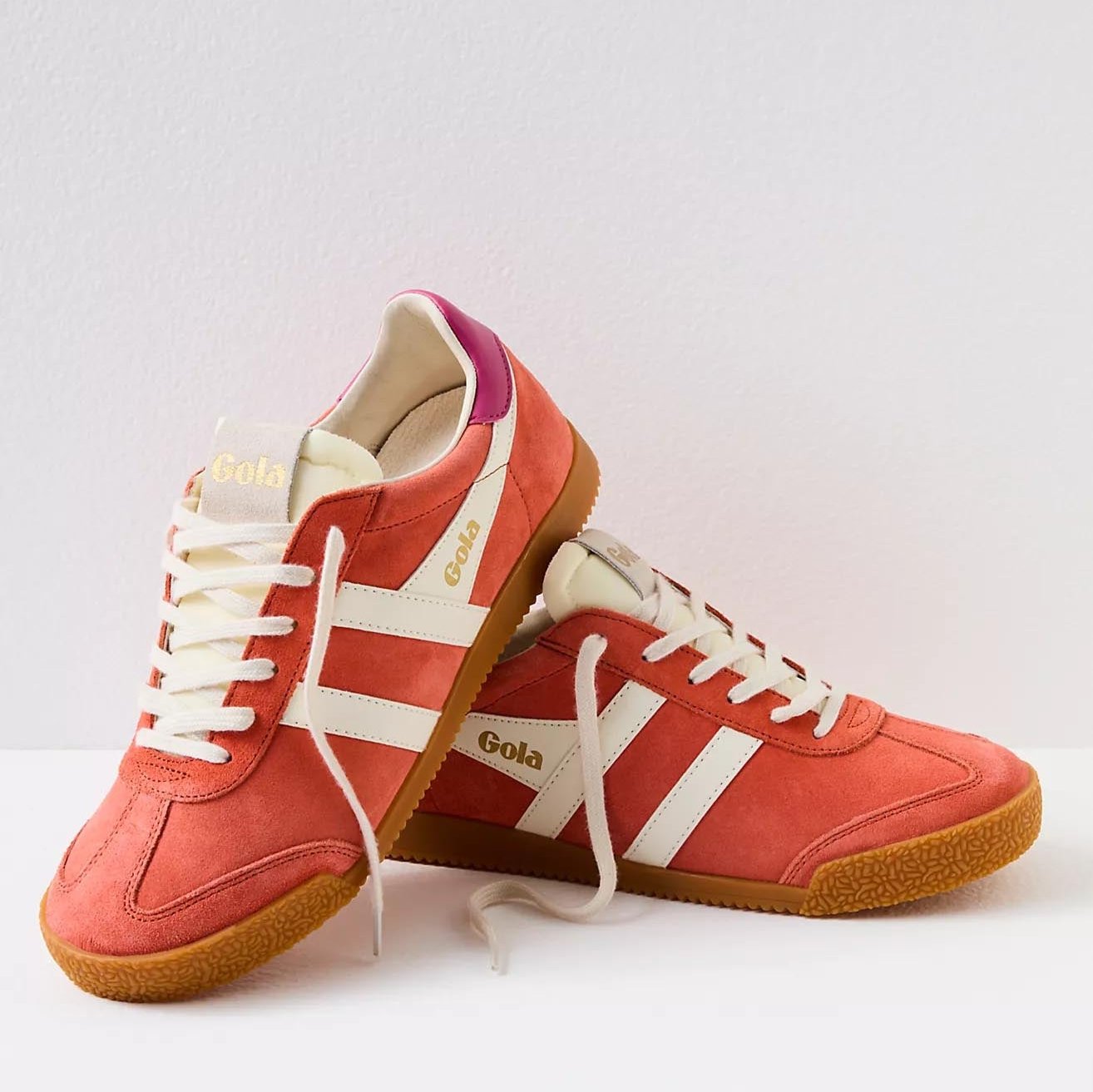 Gola sneakers in a vibrant color scheme of hot coral, off-white, and fuchsia. The shoes have a classic retro design with suede material and a gum rubber sole. The brand name "Gola" is prominently displayed on the side and tongue of the sneakers in gold lettering. The laces and stripes are off-white, providing a striking contrast to the bold coral and fuchsia colors.