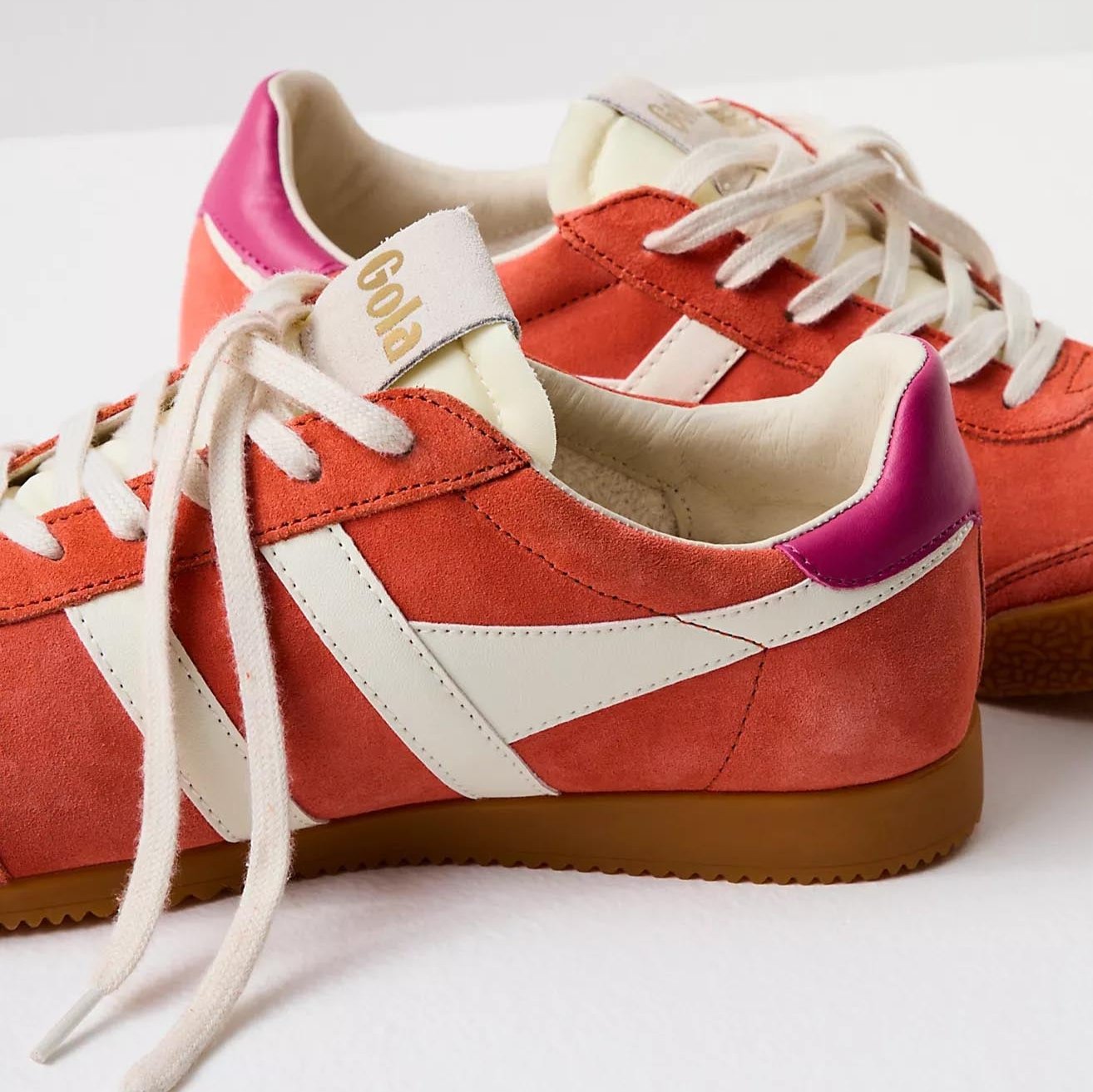 Gola sneakers in a vibrant color scheme of hot coral, off-white, and fuchsia. The shoes have a classic retro design with suede material and a gum rubber sole. The brand name "Gola" is prominently displayed on the side and tongue of the sneakers in gold lettering. The laces and stripes are off-white, providing a striking contrast to the bold coral and fuchsia colors.