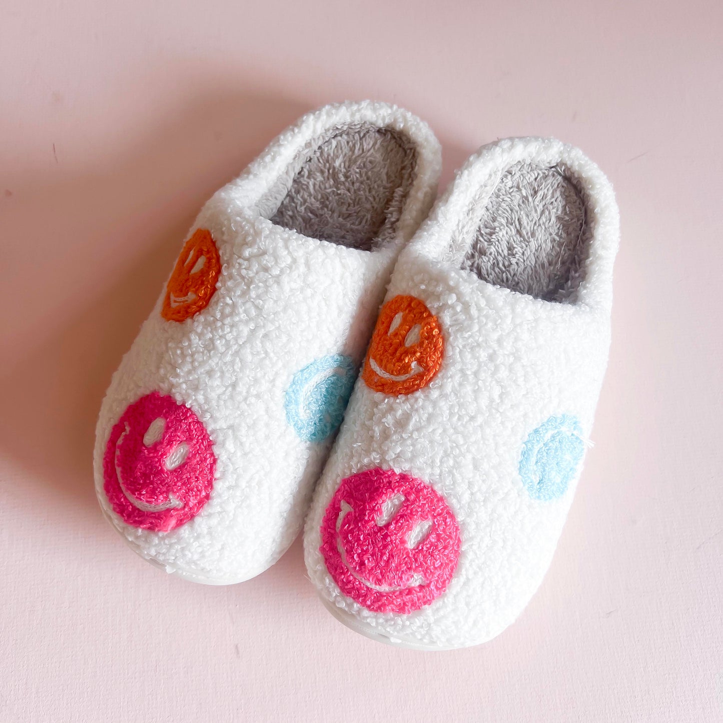Fluffy white slippers adorned with colorful embroidered smiley faces in pink, orange, and blue. These cozy and fun slippers are perfect for adding a touch of happiness and comfort to your indoor footwear collection.