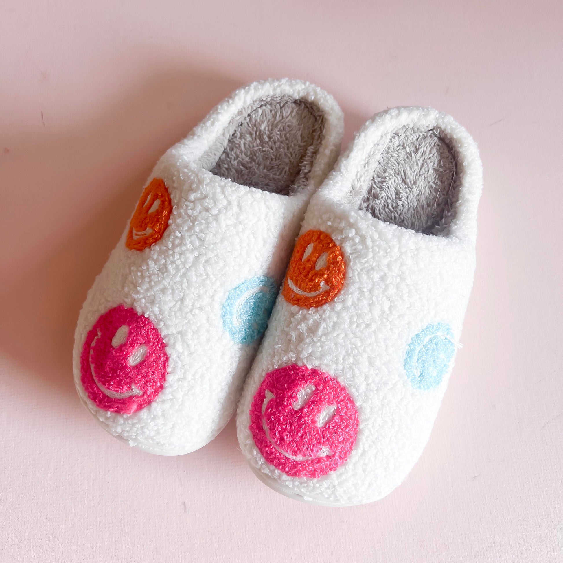 Fluffy white slippers adorned with colorful embroidered smiley faces in pink, orange, and blue. These cozy and fun slippers are perfect for adding a touch of happiness and comfort to your indoor footwear collection.