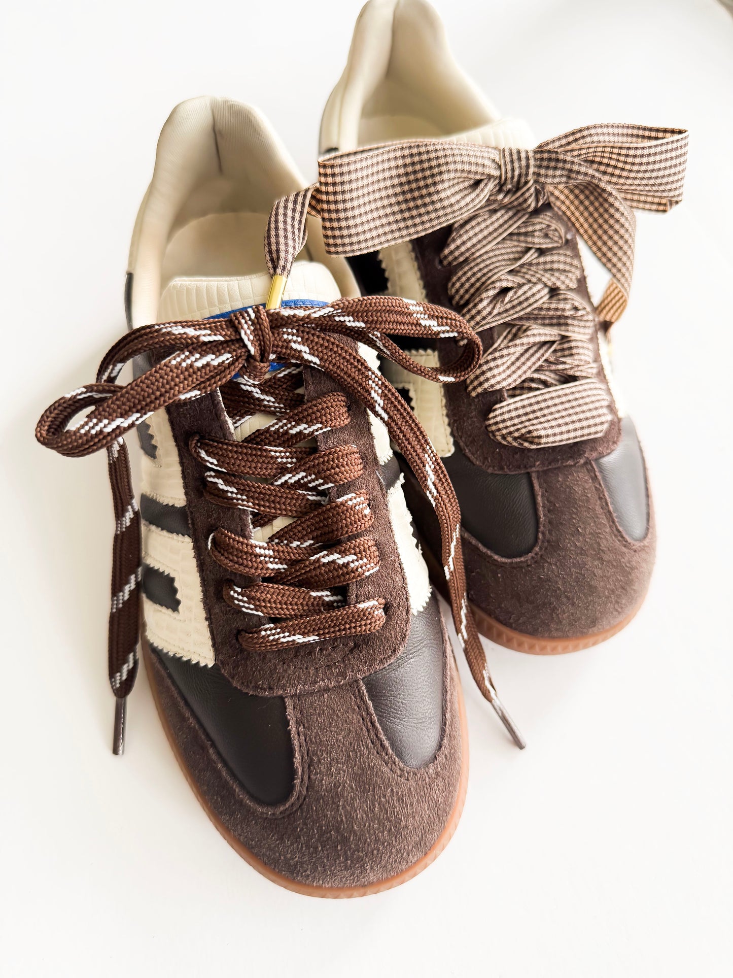 A pair of retro-style low-top sneakers in brown suede and black leather, featuring cream knit panels, patterned brown and beige laces, and oversized matching bow laces, photographed on a white background.