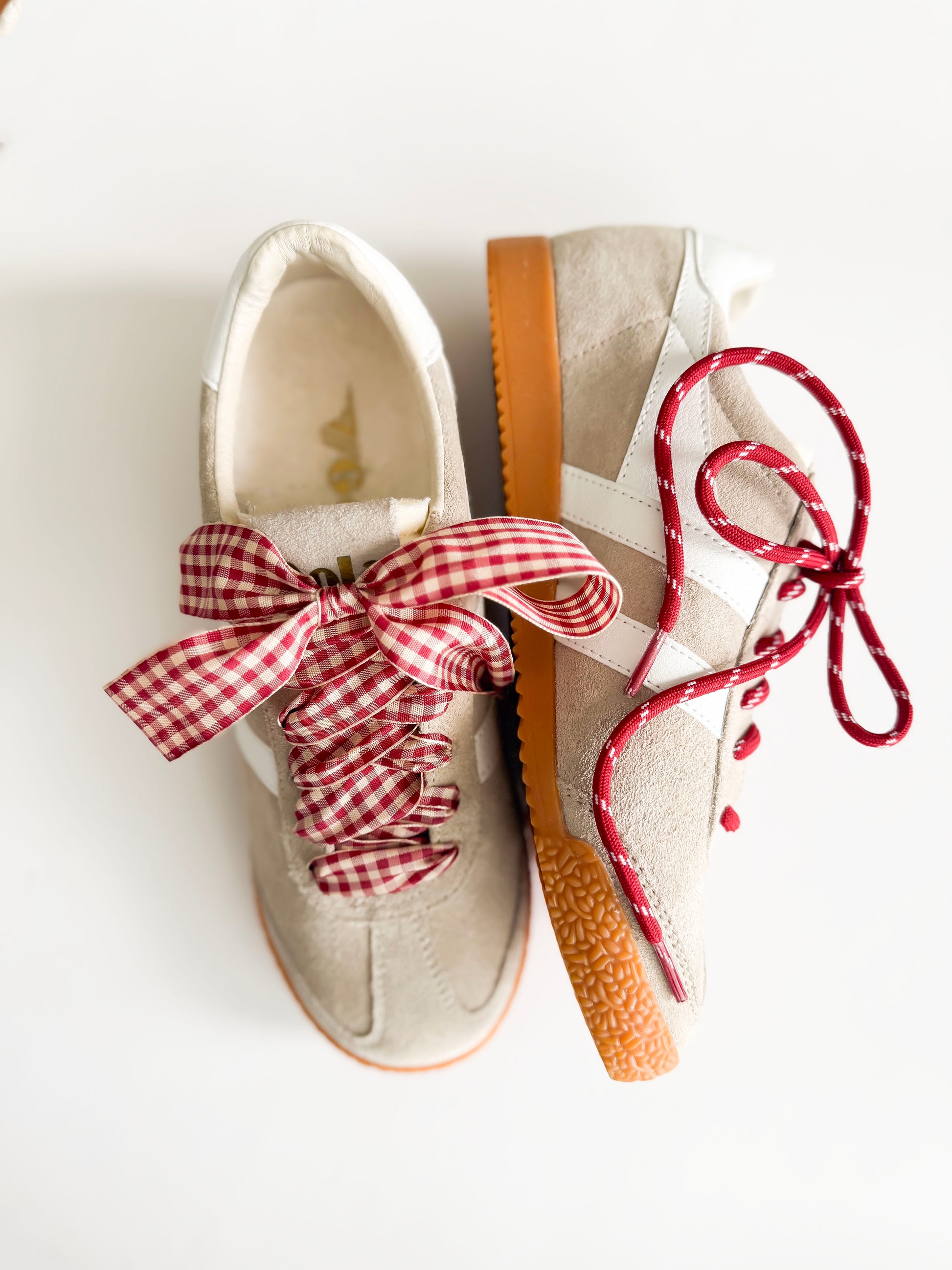 A pair of beige suede low-top sneakers with white side stripes and orange gum soles, styled with red-and-white gingham ribbon laces on a clean white background.