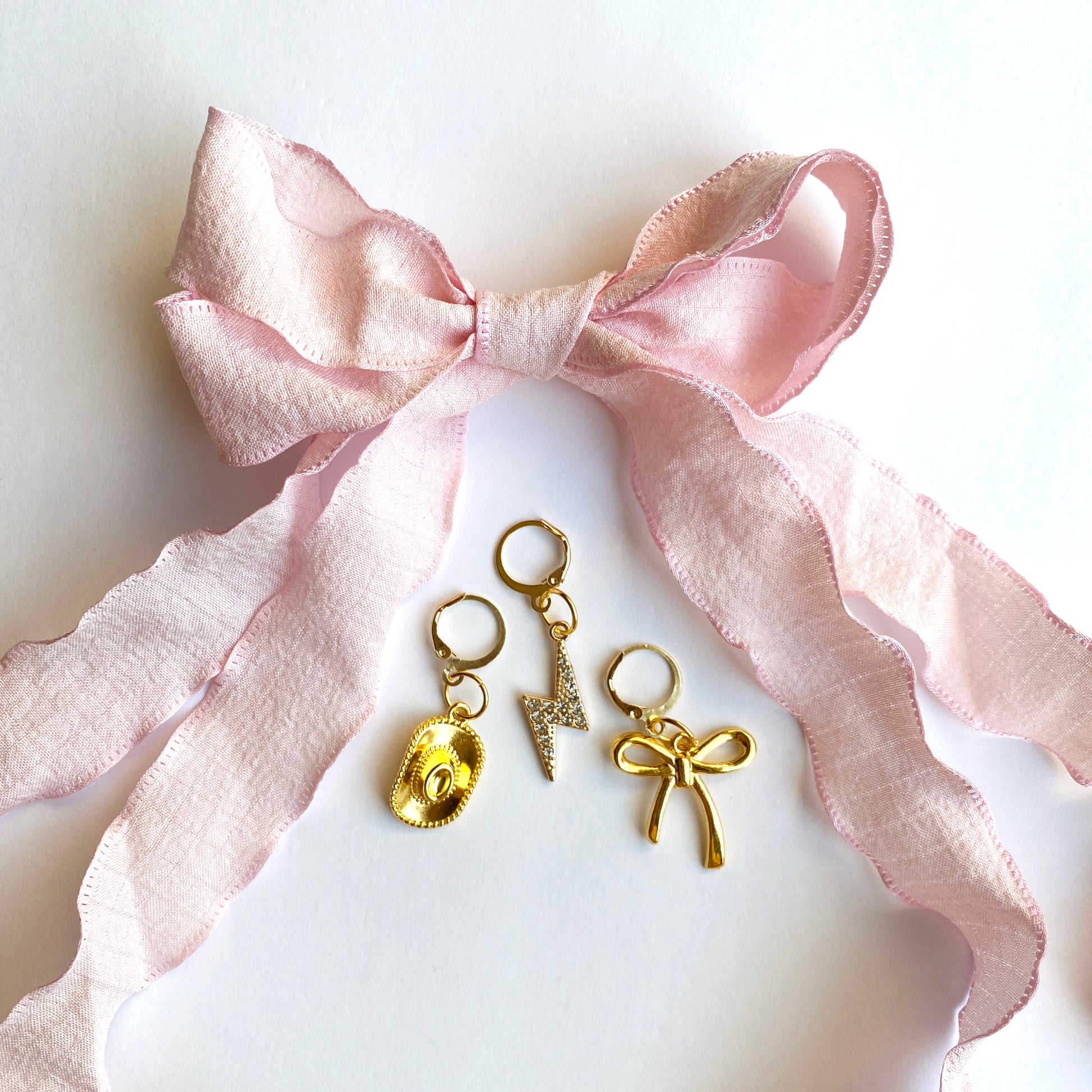 Light pink ribbon bow displayed with three gold sneaker charms — cowboy hat, lightning bolt with rhinestones, and bow — on a white background.