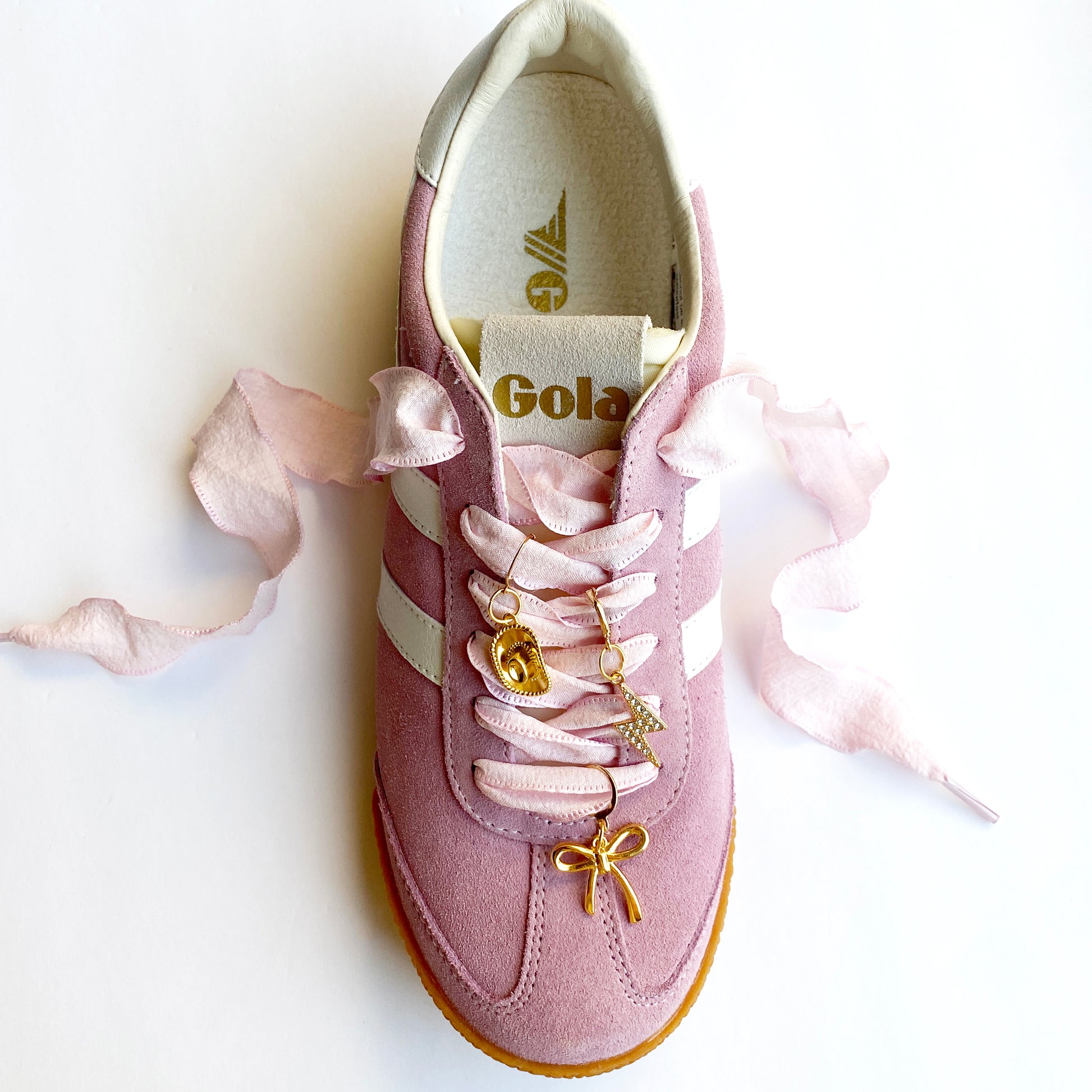 Light pink ribbon bow displayed with three gold sneaker charms — cowboy hat, lightning bolt with rhinestones, and bow — on a white background.