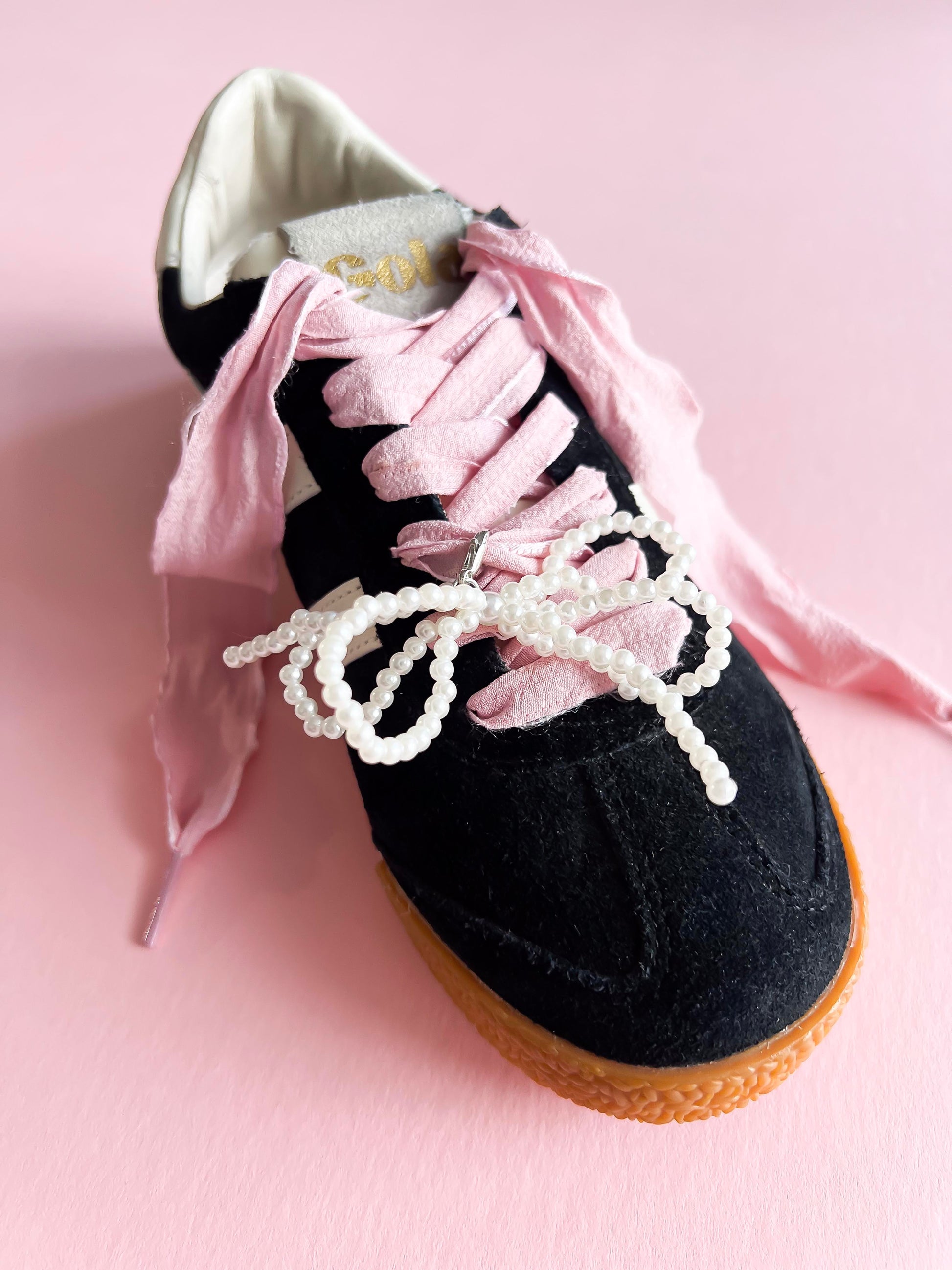 Black sneaker decorated with pink ribbon laces and a pearl bow charm, showing how the lace charm set enhances the shoe with elegant, custom detailing