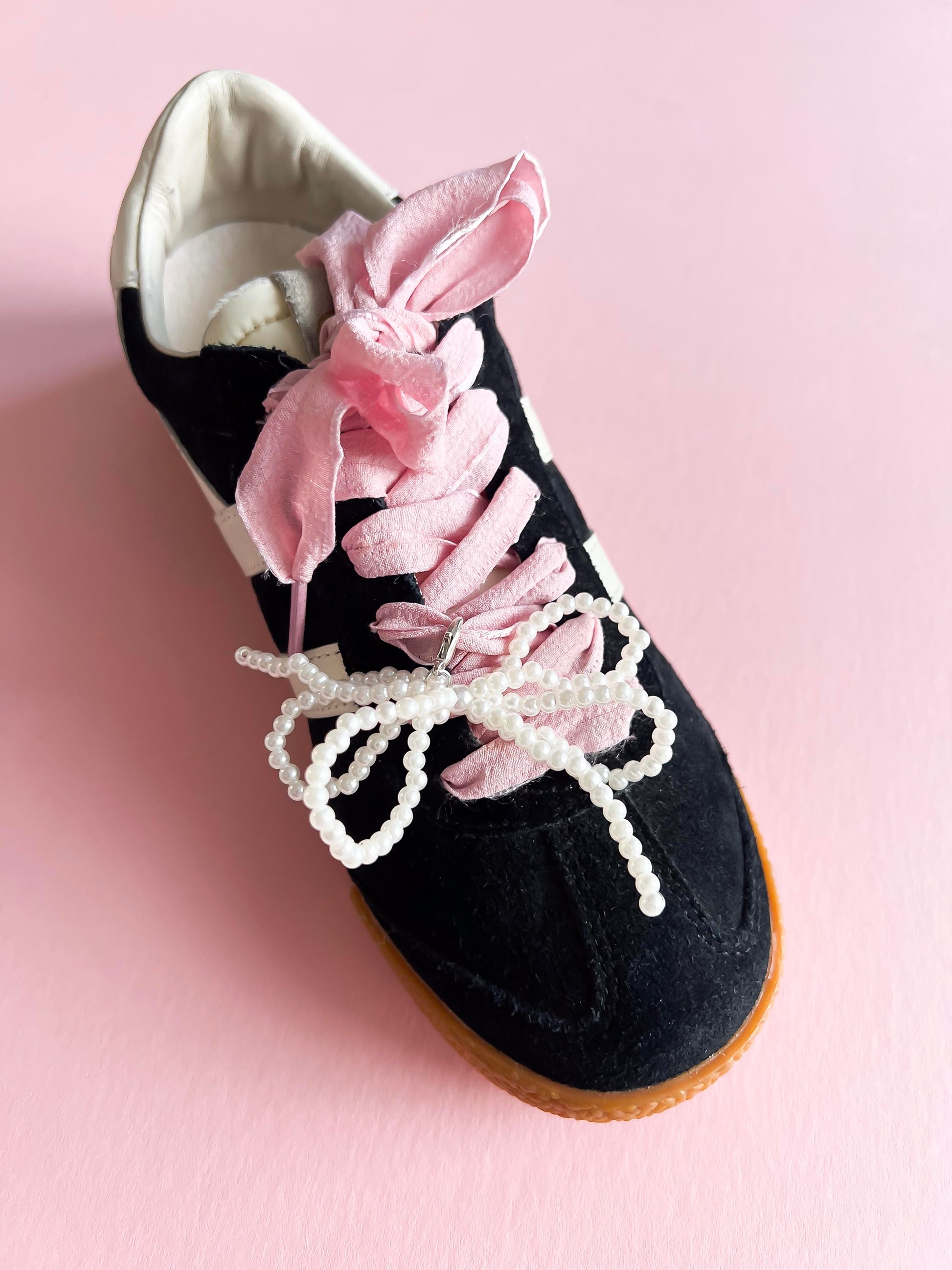 Black sneaker decorated with pink ribbon laces and a pearl bow charm, showing how the lace charm set enhances the shoe with elegant, custom detailing